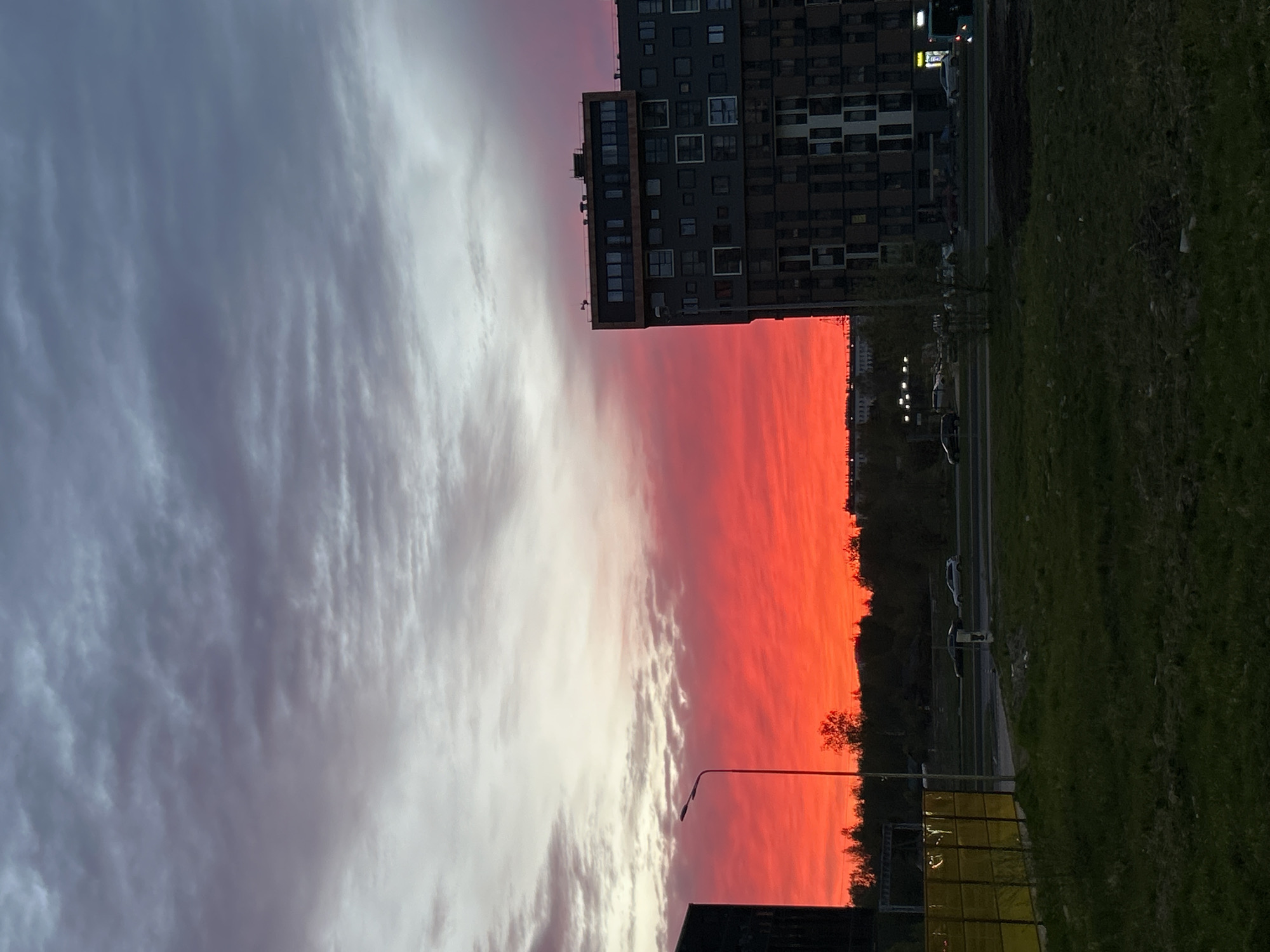 The image shows a sunset in Saint Petersburg, Russia, with a vibrant red sky dominating the center of the frame. To the left, striated white and gray clouds fill the sky. A tall, dark gray building with numerous windows stands to the upper right. Below it, a road with a few cars is visible. Green grass fills the lower right corner. A street light pole is present at the bottom center. The general environment appears to be an urban area.