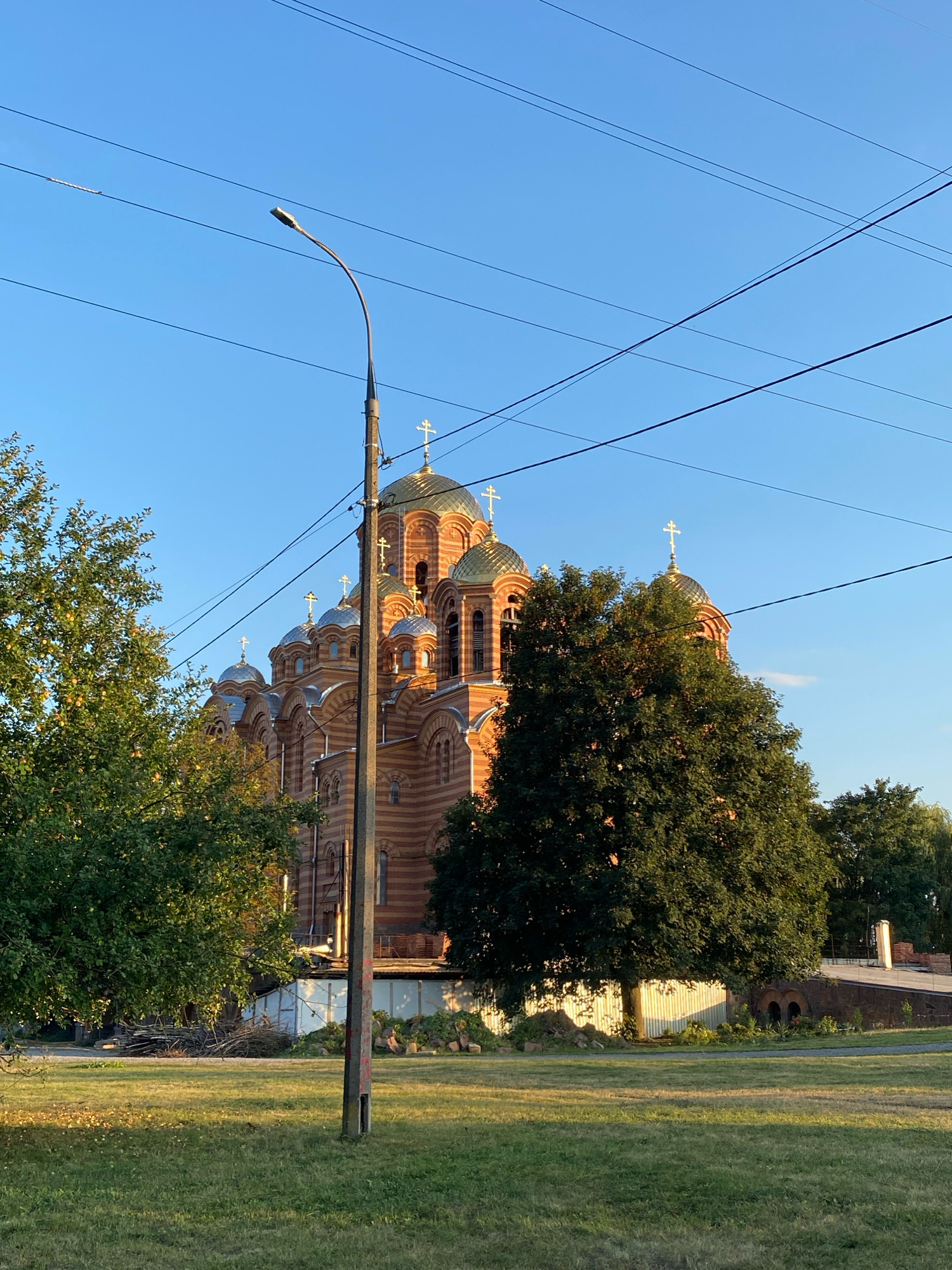 The image shows the Church of the Transfiguration in Vinnytsia, Ukraine, on a clear, sunny day. The church, made of red brick with a golden dome, is partially obscured by trees and power lines. In the foreground, a grassy field with a lamppost and various piles of debris is visible. The sky is a bright blue.