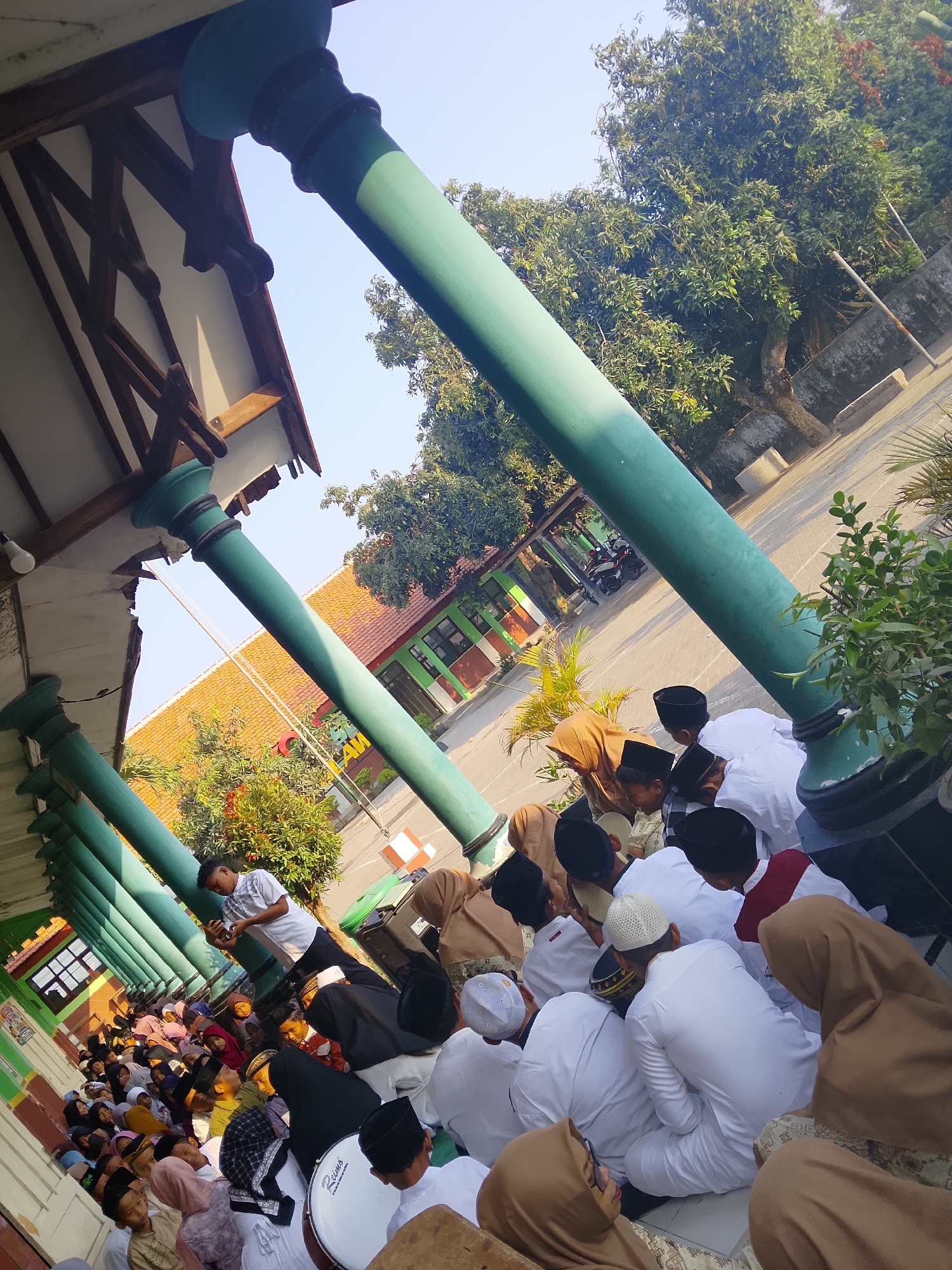 The image depicts a gathering of people, many of whom appear to be children or young adults, in an outdoor setting that resembles a school courtyard or a religious institution. The architecture suggests a building with a covered walkway supported by large, green, cylindrical columns. The ground is paved, and a building with an orange tiled roof and green and brown walls is visible in the background. Trees with lush foliage are present, and the sky is clear and blue, indicating daylight.
Several individuals are seated on the ground, many wearing white attire and black or white caps (peci), which are common in Islamic contexts in Indonesia. Some are looking down, possibly in prayer or listening to an instruction. A few adults are also present, including individuals wearing headscarves. One young man in a light-colored shirt stands and appears to be looking at his phone. A drum with "Presto" written on it is visible in the foreground, suggesting a possible musical or ceremonial aspect to the gathering. The overall atmosphere seems peaceful and organized. The provided context mentions the location as "Kepuh, Indonesia," which likely refers to a village or a specific area within Indonesia.