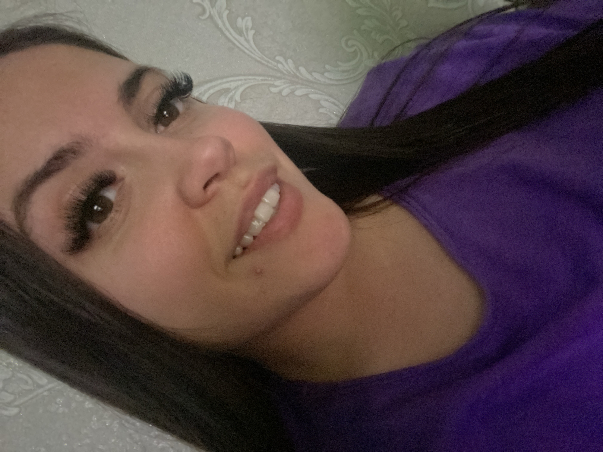 A woman with long dark hair and false eyelashes is looking at the camera and smiling. She is wearing a purple shirt. The background is a white wall with a floral pattern. The location is likely an interior setting. The city is not mentioned. There is no way to know if this image was taken in Chaikovsky, Russia.