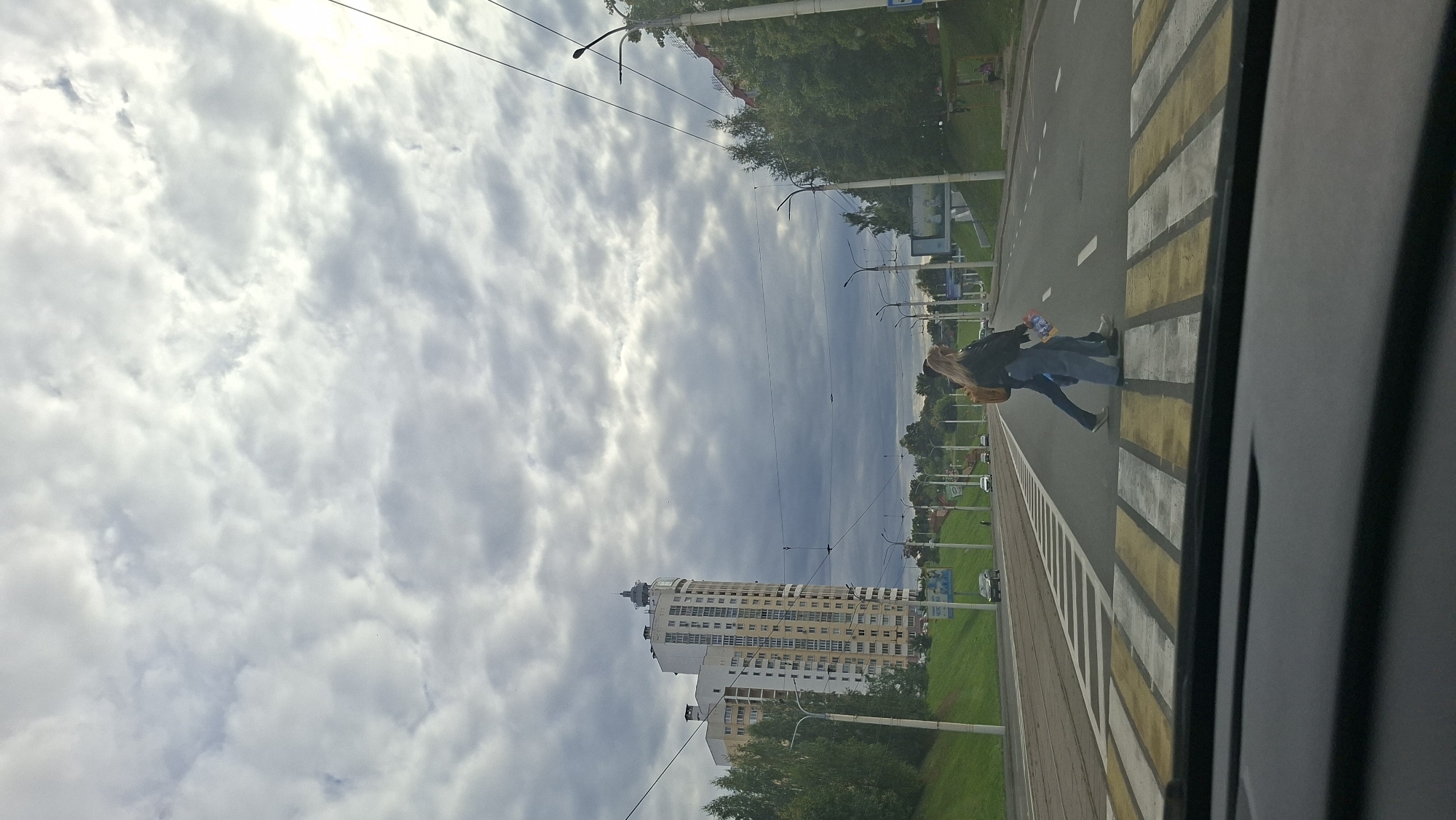 The image depicts a street scene in Vitsebsk, Belarus, under a cloudy sky. In the foreground, a pedestrian crossing with yellow and white stripes is visible, viewed from the interior of a vehicle. Two individuals, a woman with blonde hair in a black jacket and blue jeans, and a person in a brown backpack and dark pants, are crossing the street on the pedestrian crossing from right to left. The woman is carrying a light-colored bag. In the background, a tall, multi-story building with a distinctive spire dominates the skyline on the left side of the image. The building is light-colored with multiple windows and balconies. A green, grassy embankment slopes up towards the building. Streetlights and overhead wires are present along the road. Several cars are visible further down the road. Trees and foliage line the sides of the street. The overall atmosphere is overcast and urban.