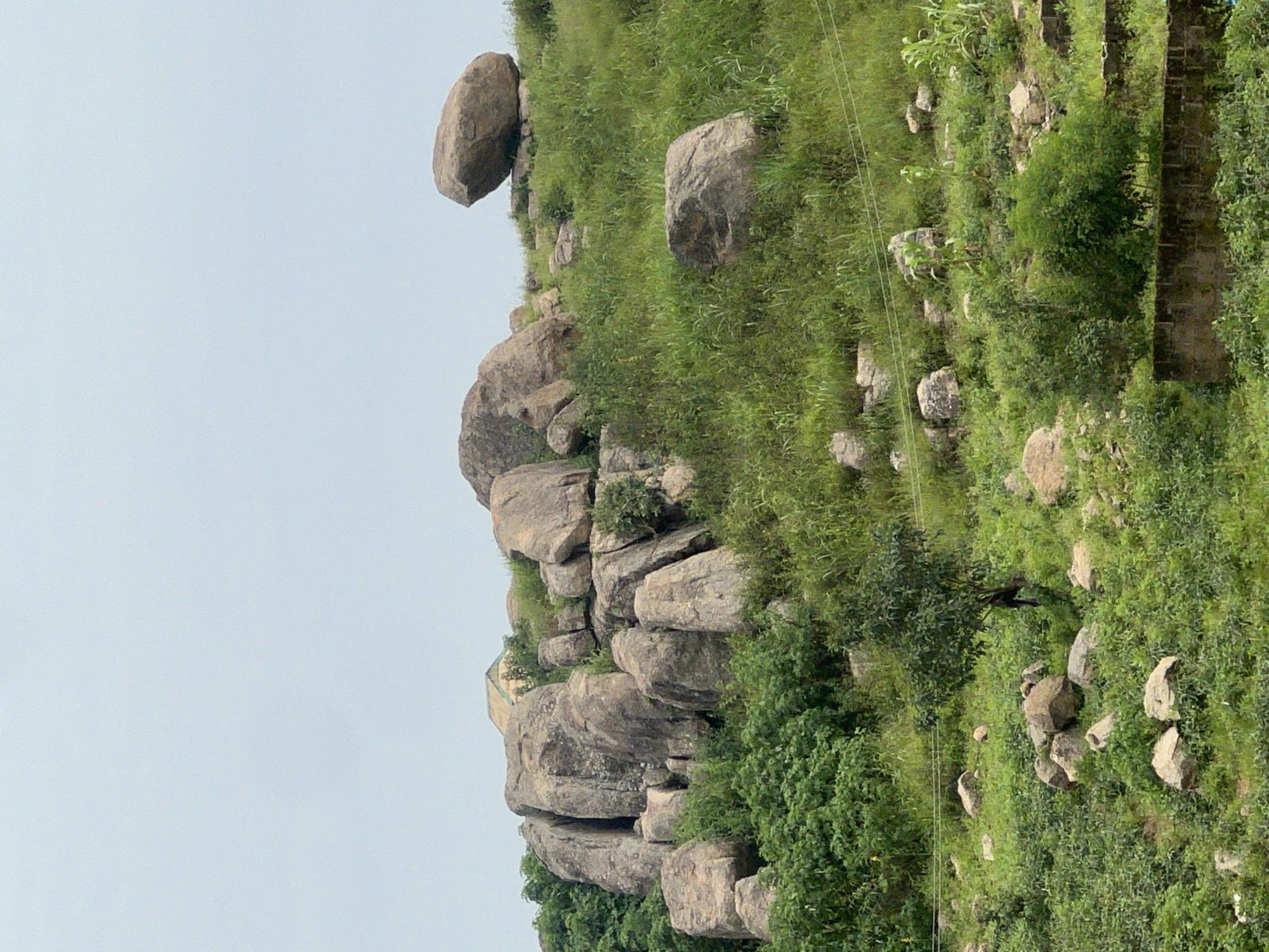 The image depicts a hilly landscape in the city of Dong, Nigeria, characterized by large rock formations scattered across a verdant terrain. The rocks are prominent features, varying in size and shape, with some clustered together to form miniature cliffs and others resting individually on the grassy slopes. Lush green vegetation, including trees and tall grasses, covers the hills, creating a natural and somewhat wild environment. In the background, a building with a light-colored roof is partially visible, suggesting human presence or settlement within the area. The sky is overcast, contributing to a soft, diffused light that illuminates the scene. The overall impression is one of a natural landscape dotted with geological features and hints of habitation.