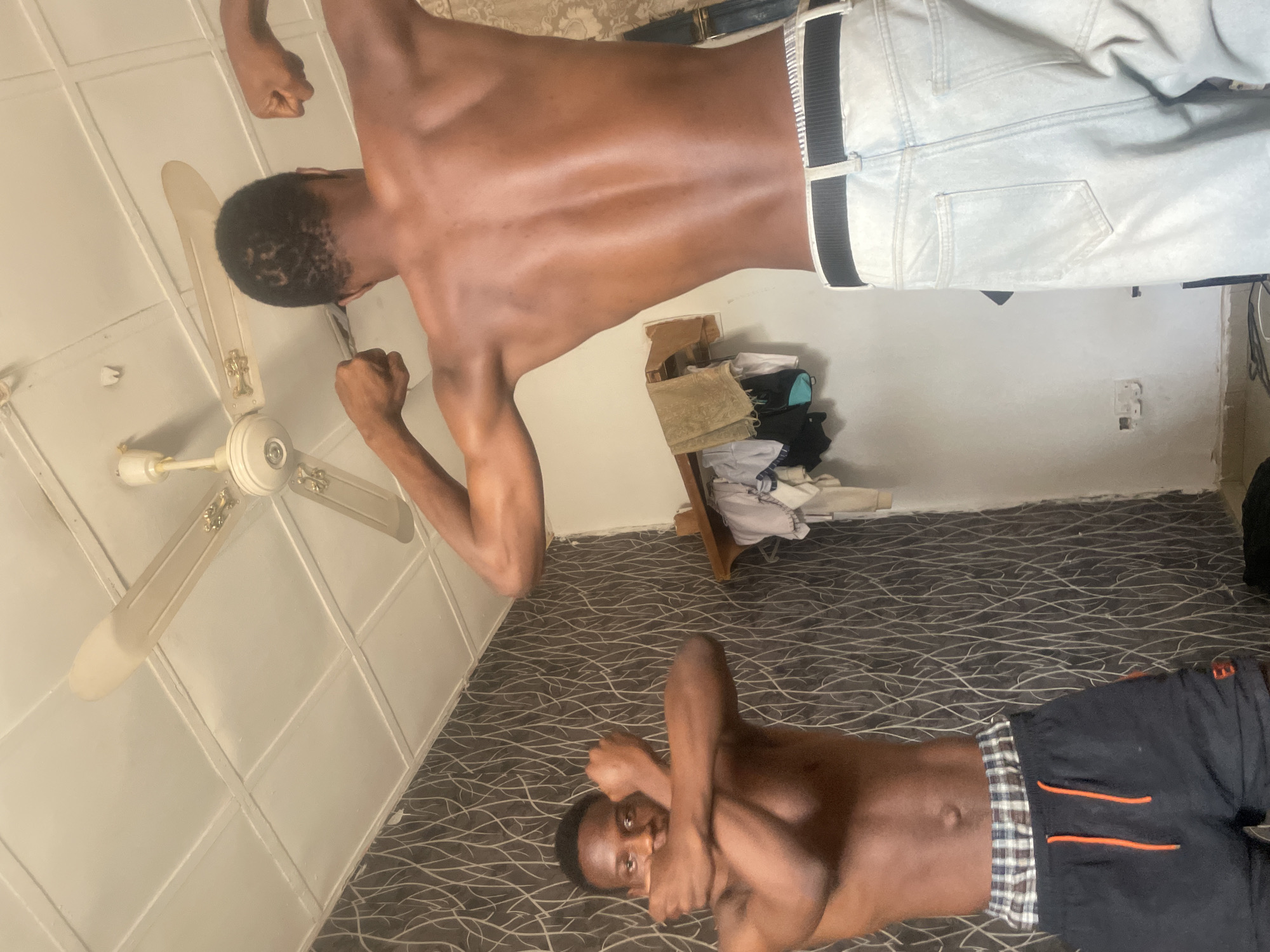 This image, captured indoors in Bauchi, Nigeria, features two young, dark-skinned males posing. The photograph is rotated 90 degrees clockwise from its intended orientation.
One male is positioned as if standing, shirtless and wearing light-colored pants, flexing his biceps with both arms raised. His back is largely towards the viewer, with his head slightly turned. Above him, a white ceiling fan with four blades and golden accents is mounted on a white, panelled ceiling.
The second male is lying on a dark floor covered with a patterned carpet or rug featuring wavy, light-colored lines. He is also shirtless, wearing dark shorts with orange stripes and a plaid waistband. He looks directly at the camera with his arms crossed over his chest in a casual, possibly playful, pose.
In the background, a light-colored wall is visible, against which stands a wooden shelf or small piece of furniture laden with folded clothes. A white electrical outlet is also discernible on the lower part of the wall. The scene suggests a relaxed, candid, or playful moment taking place within a personal indoor setting, likely a room in a home. No text is visible in the image.