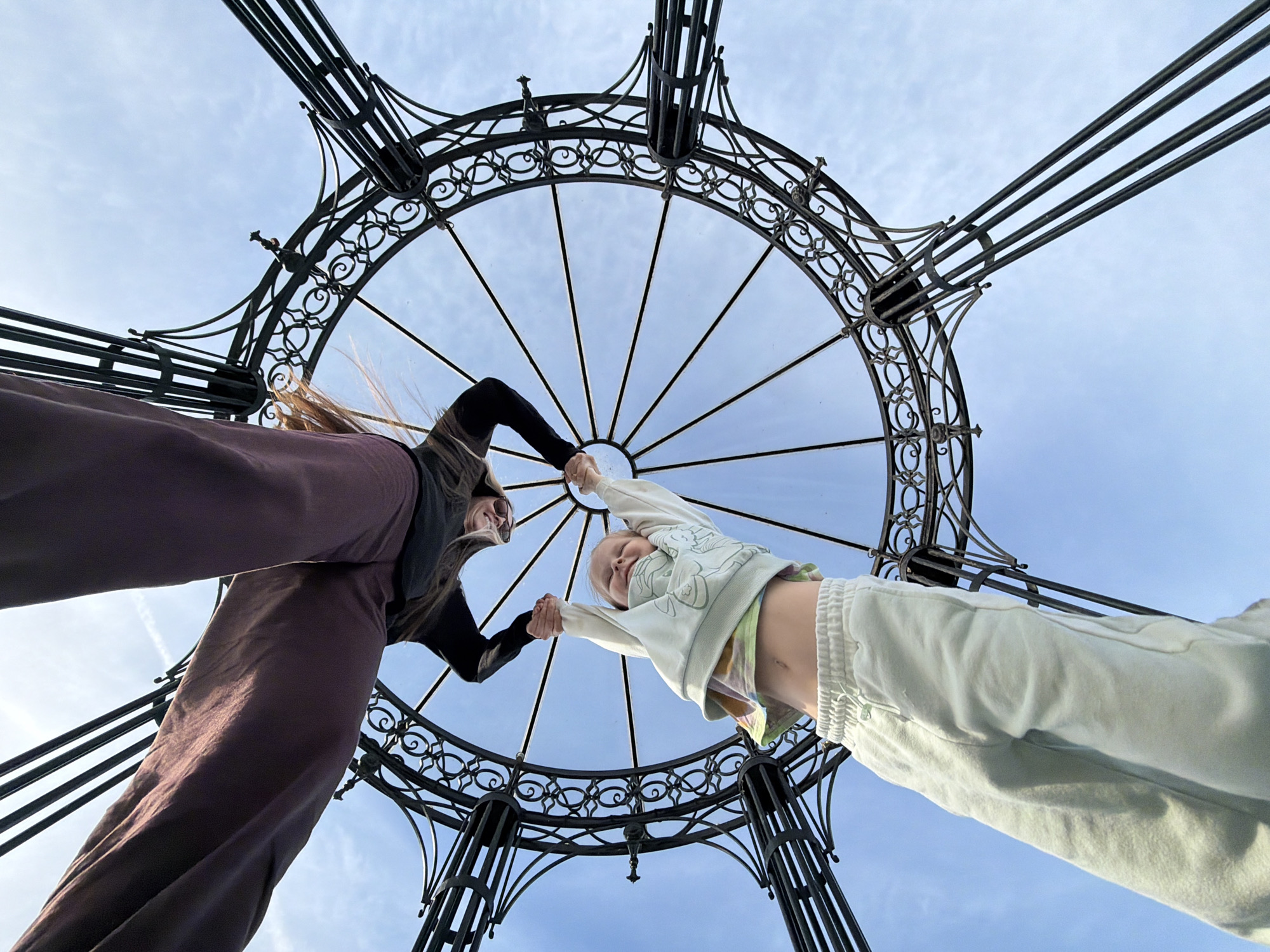 Two individuals, one appearing to be an adult and the other a child, are holding hands beneath an ornamental metal structure. The adult is on the left, wearing dark-colored pants and a dark top, while the child is on the right, wearing light-colored pants and a light-colored top. The photo is taken from a low angle, looking up at the structure and the people beneath it. The metal structure has a circular, open design with radiating lines and ornate detailing. The background is a light blue sky with wisps of clouds. This location is in the city of Kamyshin, Russia.