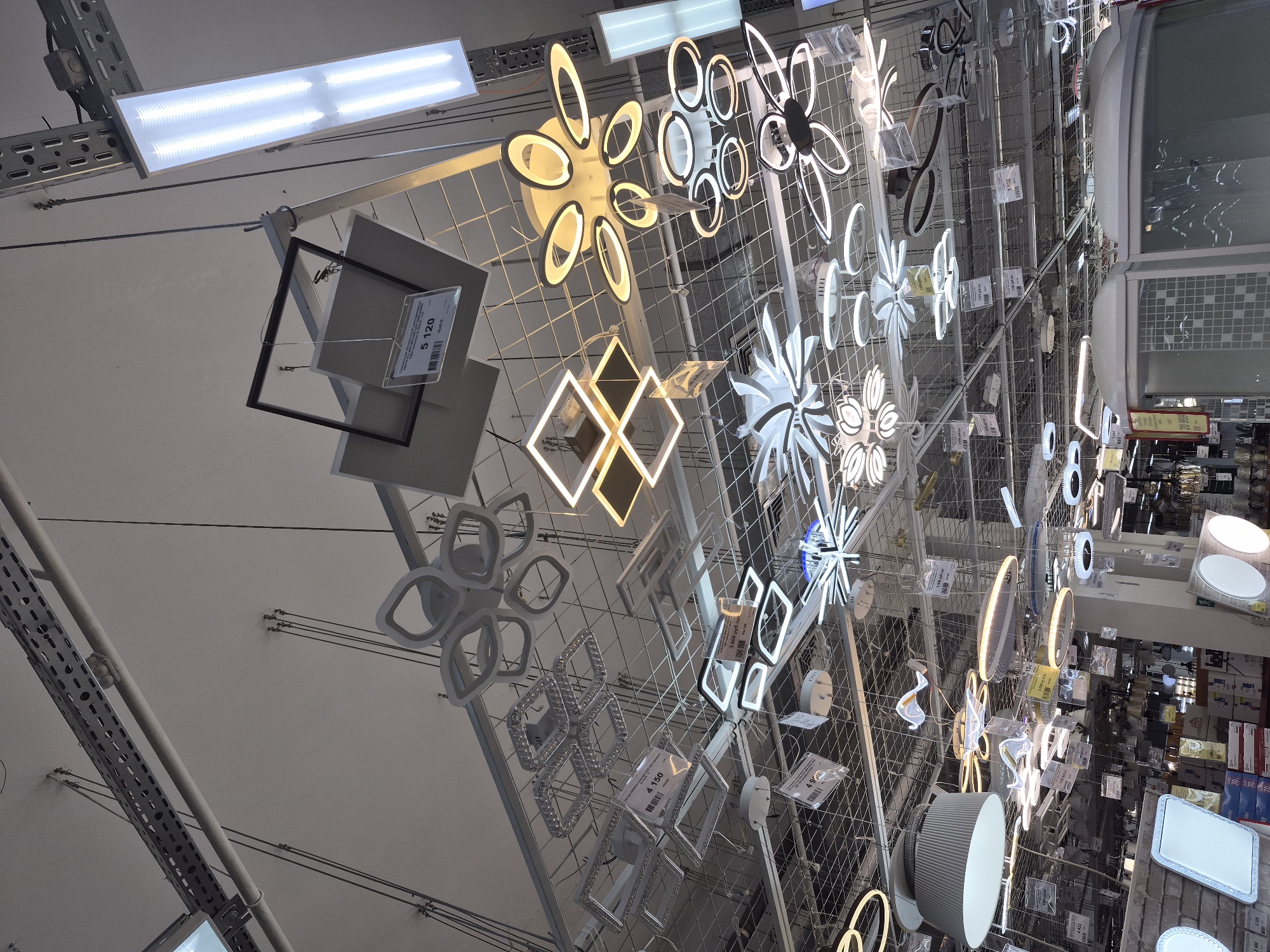 The image shows a display of modern light fixtures hanging from a metal grid in a retail setting. Various styles are visible, including flower-shaped LED lights, geometric designs, and circular fixtures. Each light is accompanied by a price tag. The setting appears to be a lighting section within a larger store, potentially in Tobolsk, Russia. The image is taken from a low angle, looking up at the display.