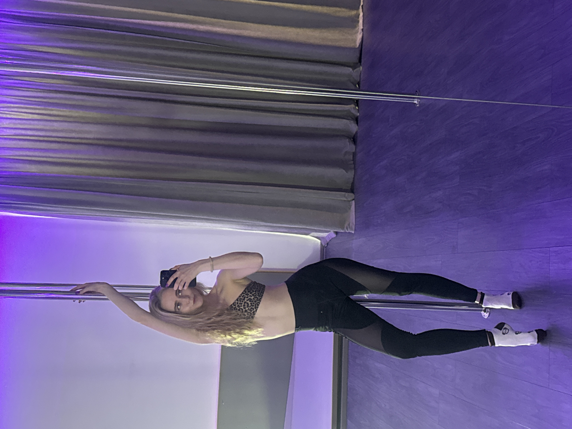A young woman with long, wavy blonde hair is captured in an indoor setting, likely a pole fitness studio or gym. She is wearing a leopard print sports bra and black leggings with sheer mesh panels on the thighs and calves, complemented by white socks with black details. She holds a black smartphone in her right hand, taking a selfie, while her left arm is extended and rests gracefully on a chrome pole. Another parallel chrome pole is visible in the background, both extending vertically.
The room is dimly lit with a dominant purple hue, which casts a vivid glow on the dark, possibly wooden or laminate, floor and subtly illuminates the woman. The background features a wall covered with dark grey, horizontally draped fabric, resembling curtains or acoustic panels, which adds texture to the scene. The woman maintains a calm and focused expression as she looks towards her phone, striking an athletic and dynamic pose that suggests movement or a specific pole fitness maneuver. The overall atmosphere is artistic and energetic, defined by the dramatic lighting and the woman's confident pose. No visible text is discernible in the image.