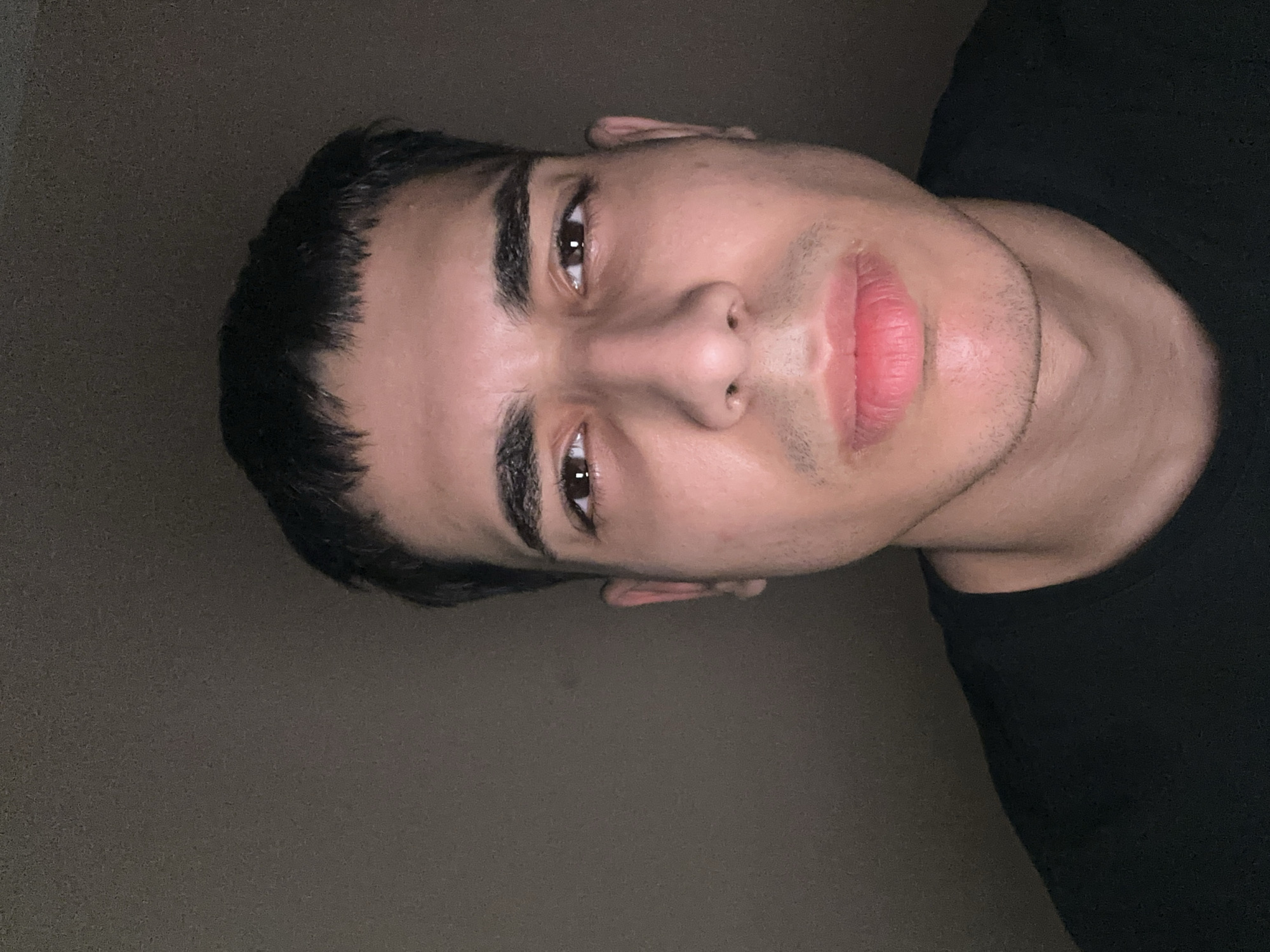 A close-up portrait of a light-skinned male with dark hair and eyebrows. The subject is facing the camera, with the image rotated slightly. His expression is neutral. He is wearing a black shirt. The background is a plain, dark wall. There is no indication of location in Kozloduy, Bulgaria.