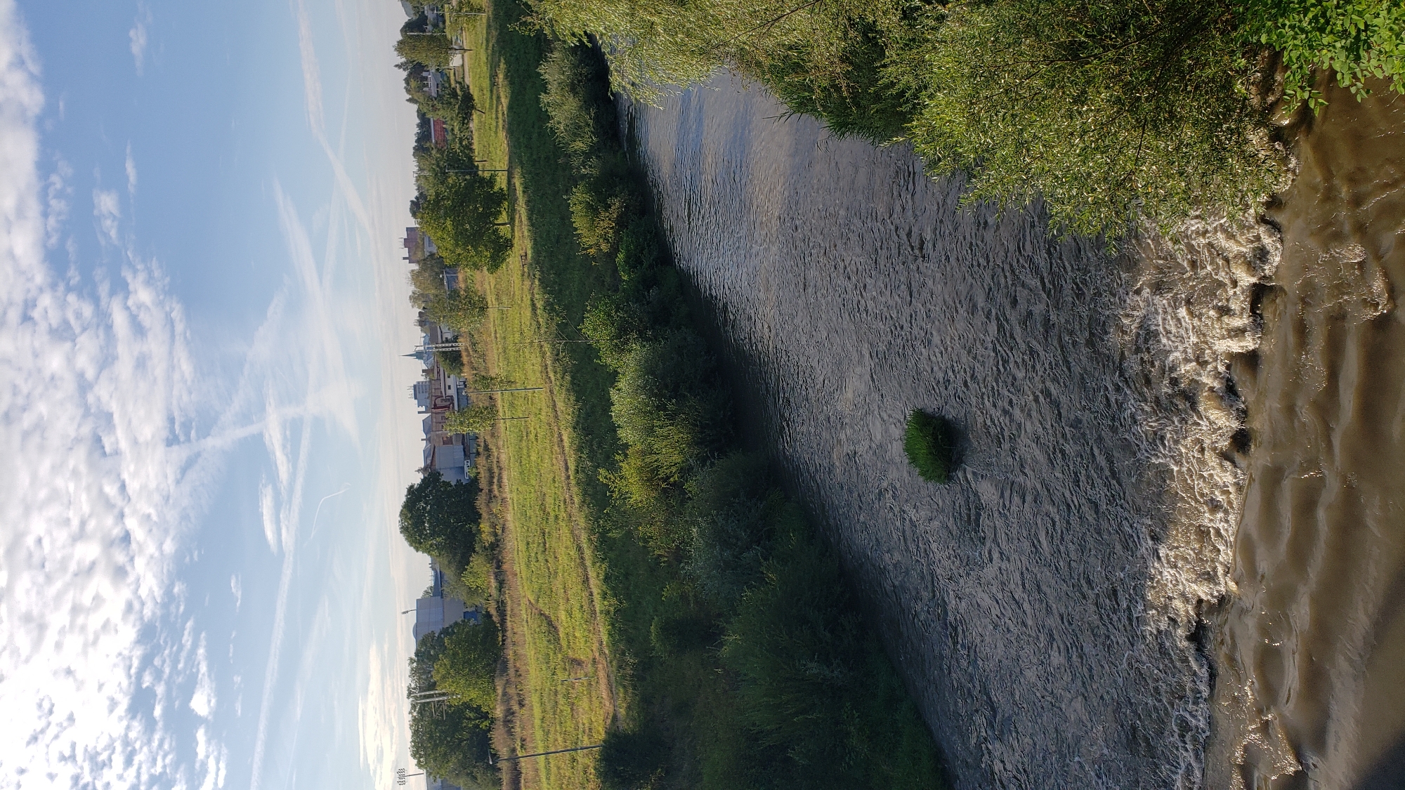 The image captures a serene view of the Torysa River in Prešov, Slovakia. The river flows gently, with a small patch of grass in the middle. The banks are lined with lush green vegetation and trees. In the background, a cityscape with buildings and trees can be seen under a clear blue sky with scattered clouds and contrails. The overall scene depicts a tranquil natural setting within an urban environment.