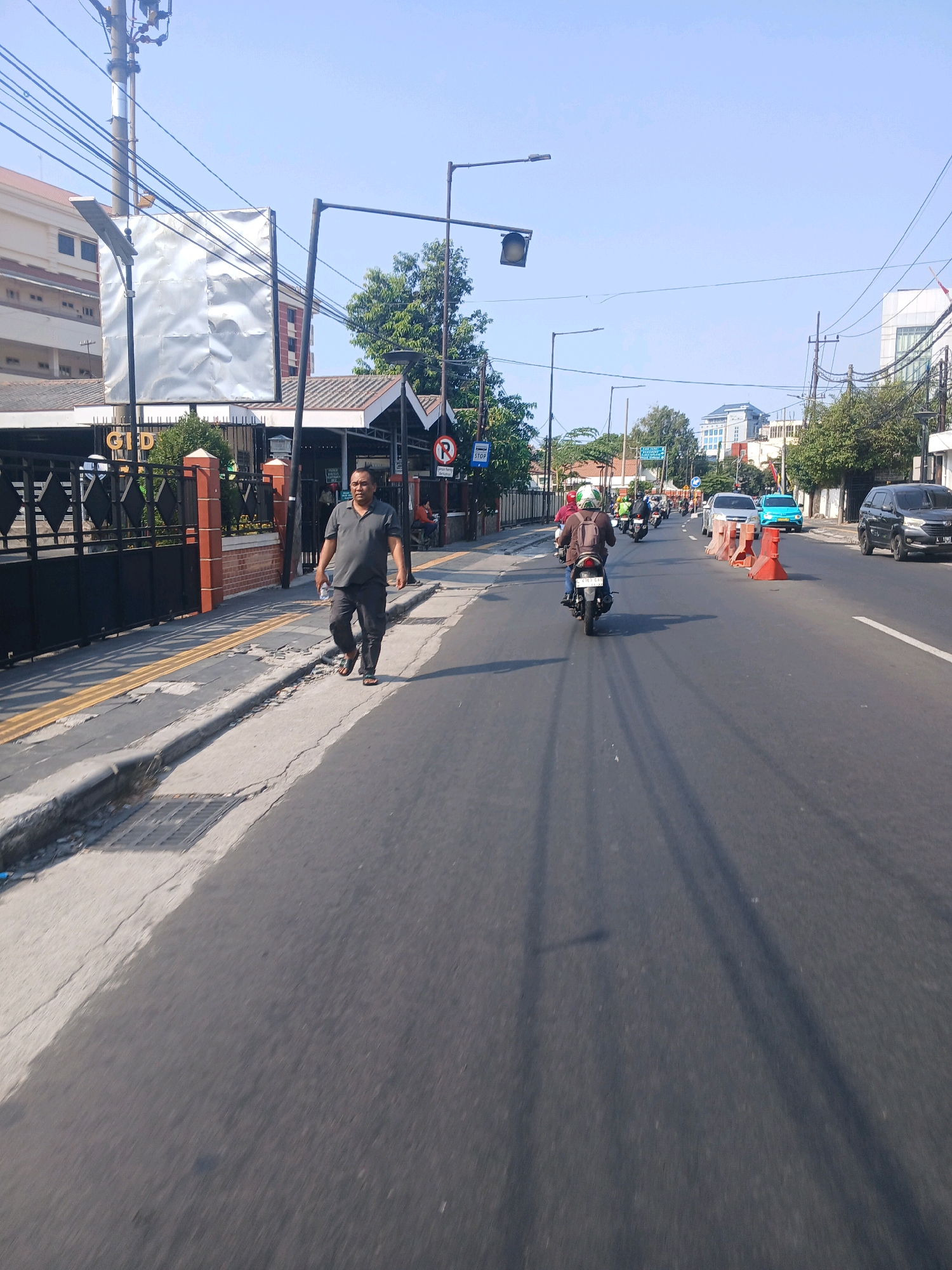 A man in a gray polo shirt and dark pants walks along a sidewalk on a street in Surabaya, Indonesia. The street is paved and has multiple lanes of traffic. Several motorcycles and cars are visible on the road. In the background, there are buildings, trees, and a clear blue sky. A traffic light and street lamps are also present. The sidewalk has a yellow tactile paving for the visually impaired. A fence with black metal bars and red brick pillars borders the property on the left. A large, metallic billboard is attached to a pole on the left side of the street.