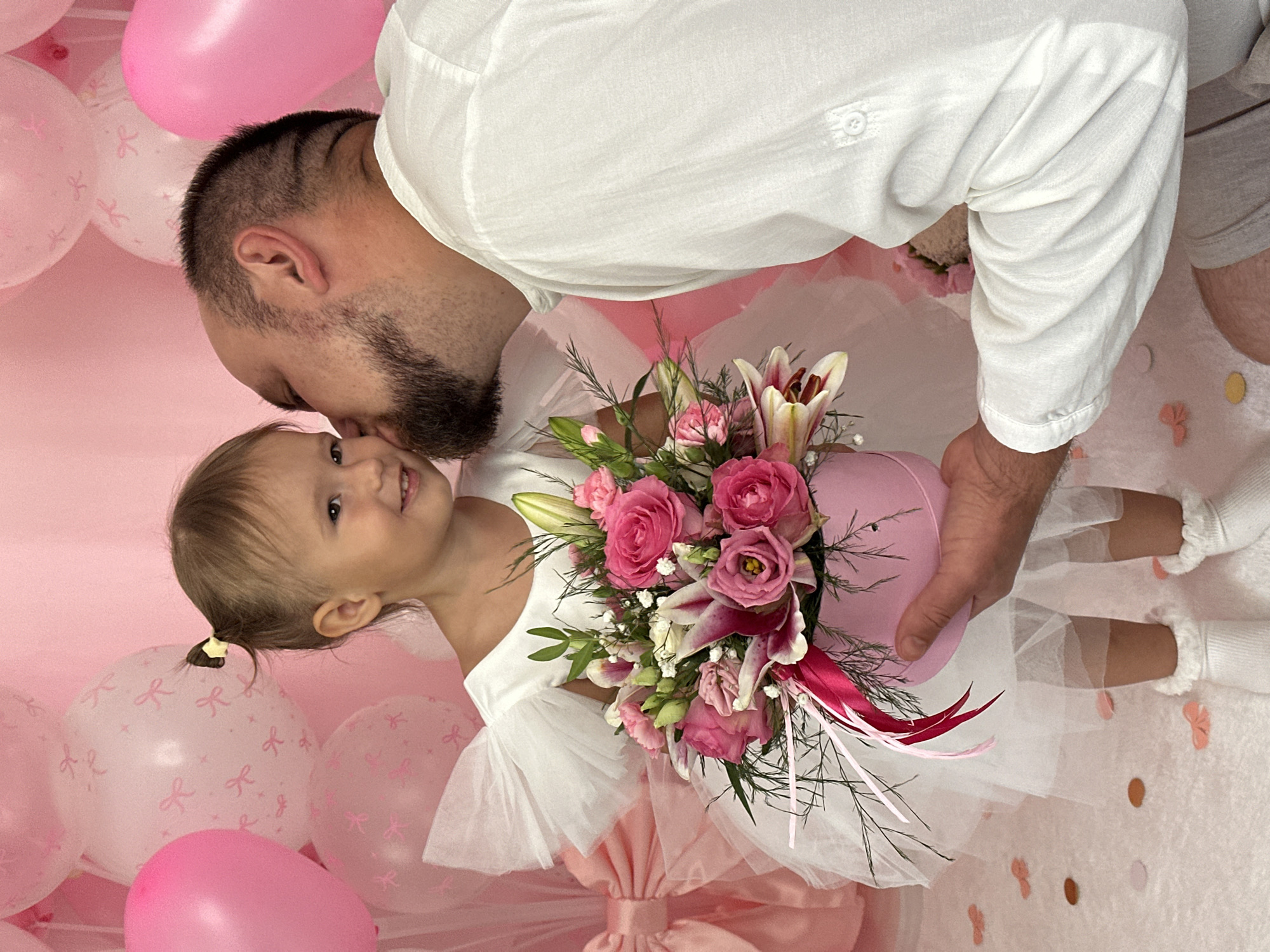 A man wearing a white shirt kisses a young girl on her cheek. The girl is wearing a white dress and holding a pink box of flowers. She has a ponytail and is smiling. In the background, there are pink balloons and a pink curtain. The scene appears to be a celebration or special occasion.