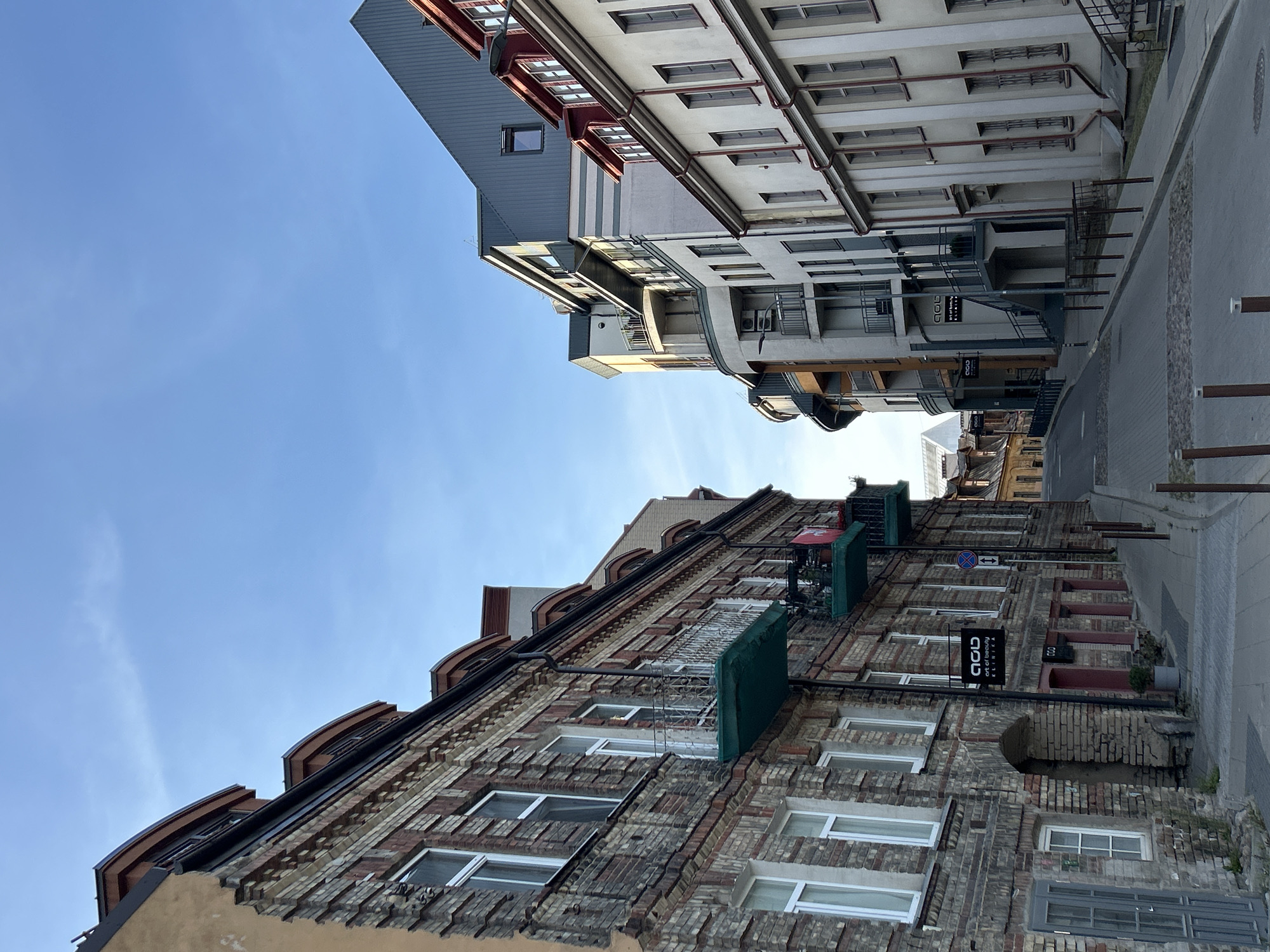 The image captures a narrow street in the Old Town of Vilnius, Lithuania, under a clear, bright blue sky. The street slopes gently upwards and curves to the right, lined with buildings of varying architectural styles and ages.
On the left, an old, weathered brick building dominates the foreground. Its facade features a mix of light and dark bricks, with a slightly damaged plaster section near the top left corner. Several windows with white frames are visible, as are two balconies with green awnings and white railings. One balcony has plants and a small flag or banner hanging from it. A dark sign with white lettering, "GOU ART OF BEAUTY," is mounted on the brick wall. An arched entryway leads into the building.
To the right, a more modern building with a curved facade rises. This building has a white exterior with gray horizontal bands and numerous balconies with black railings. Further down the street on the right, another building with a cream-colored facade and red trim around the windows is visible. Its architecture appears older, with tall, narrow windows.
The street itself is paved with cobblestones, and a sidewalk runs along the right side. Several bollards are placed along the edge of the sidewalk. In the distance, more buildings of the Old Town can be seen, with traditional tiled roofs. The lighting suggests it is either early morning or late afternoon, with the sun casting long shadows. There are no people visible in the scene.