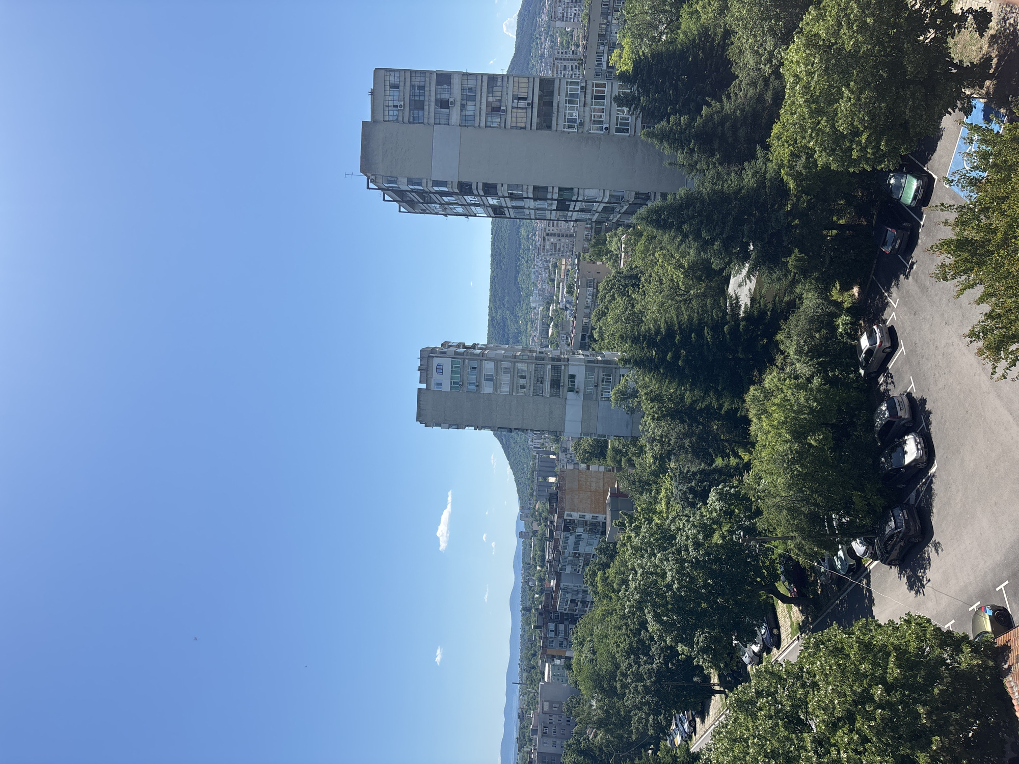 A bright, sunny day is captured in an aerial view of a city. In the foreground, a parking lot is filled with numerous cars, mostly sedans and SUVs, some parked in designated spots and others under the shade of lush green trees. Beyond the parking lot, dense foliage of various trees, including tall evergreens, dominates the mid-ground.
Further back, two tall, multi-story apartment buildings stand out. The building on the right is a light-colored, rectangular structure with many windows and balconies. To its left, another similar apartment building is visible, slightly shorter and with a different architectural style.
In the background, a sprawling urban landscape unfolds with more apartment buildings of varying heights and colors, interspersed with more greenery. Rolling hills, covered in trees and vegetation, form the distant horizon, under a clear, vibrant blue sky with a few wispy white clouds. The overall impression is a lively city scene bathed in warm sunlight.
