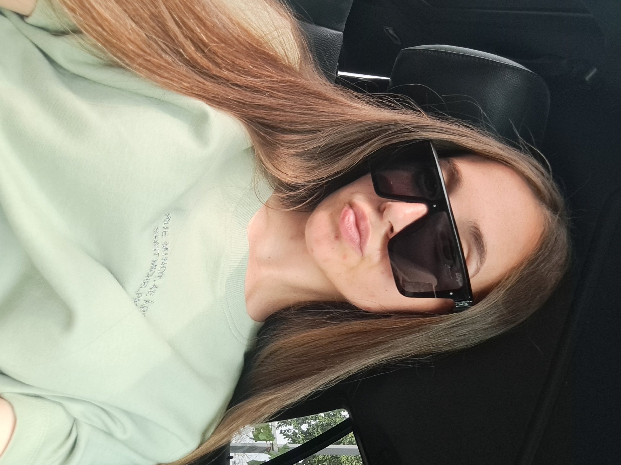 A young woman with long, light brown hair is sitting in the driver's seat of a car. She is wearing large, black, square-shaped sunglasses and a light green long-sleeved shirt with some writing on it. Her lips are slightly pursed, and she is sticking out her tongue slightly. The interior of the car, including the black leather headrest and part of the dashboard, is visible. Outside the car window, trees and foliage can be seen. The image appears to have been taken during the daytime. The scene does not provide any specific indicators of the location being Vypasne, Ukraine.