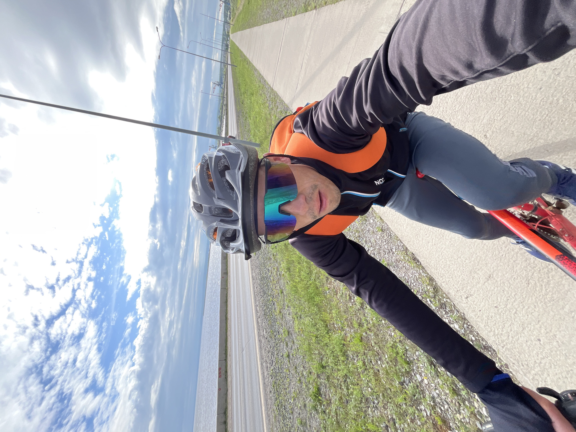 A man wearing a helmet, sunglasses, a black long-sleeved shirt with an orange vest, and grey pants is cycling on a paved path. He is looking towards the camera. The path is next to a road and a body of water, with a cloudy sky overhead. In the distance, there are streetlights and some industrial structures, possibly cranes. The scene appears to be located in the city of Chaikovsky, Russia, specifically along the Kama River embankment.