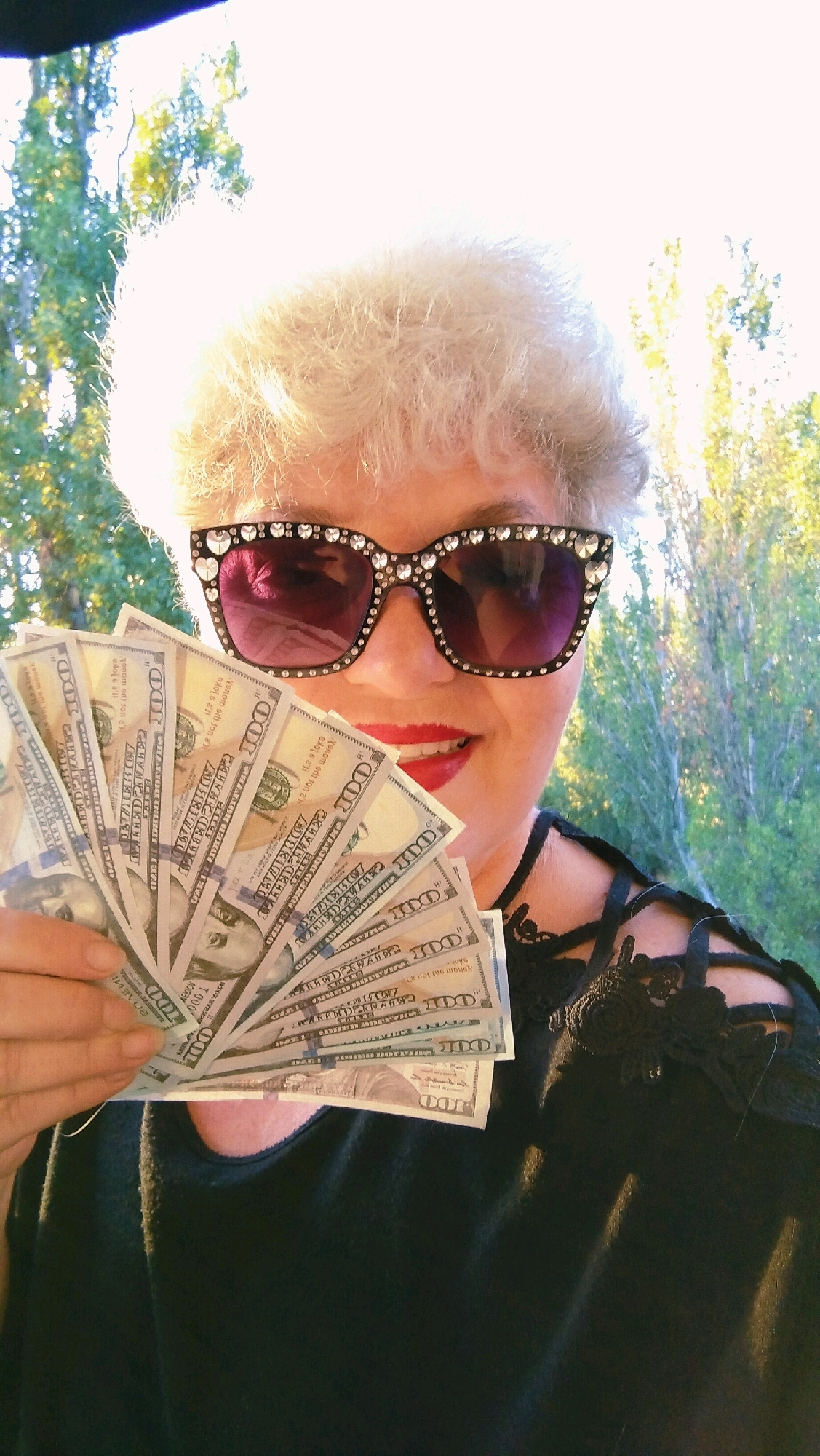A woman with short, curly, light-colored hair and red lipstick is holding a fan of US hundred-dollar bills in front of her face. She is wearing oversized, rhinestone-embellished sunglasses. The background consists of trees and a bright sky, suggesting an outdoor setting. The woman is wearing a black top with decorative straps on the shoulders. The image is a close-up portrait, with the focus on the woman and the money. There is no indication of the city of Horishni Plavni, Ukraine.