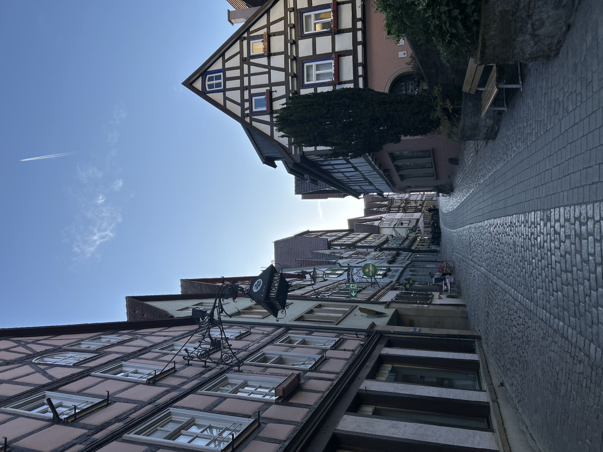 The image shows a street scene in a city. The architecture is characterized by half-timbered buildings with exposed wooden beams and white or light-colored infill. The street is paved with cobblestones and curves slightly. The buildings have multiple windows, some with shutters, and some are adorned with signs or decorations. A contrail is visible in the blue sky. The overall environment suggests a historical or traditional urban setting. The location is Unknown, Unknown.