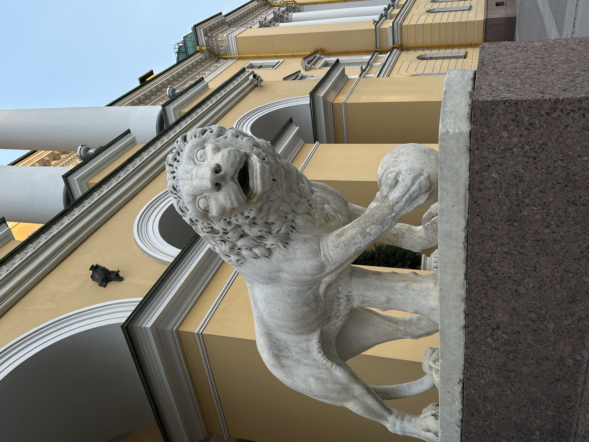 A ground-level, low-angle shot showcases a marble lion statue standing guard outside the Winter Palace in Saint Petersburg, Russia. The statue is positioned in the foreground, to the left of a two-toned plinth of grey and red granite. The statue is of a lion with its mouth wide open, with a curly mane. The lion is sculpted holding a ball. Behind the statue and plinth, the façade of the Winter Palace rises, rendered in a pale yellow hue. White architectural details outline the building's structural elements, including archways. A bronze sculpture of a turtle is visible on the palace wall. The sky is visible in the upper left corner of the frame.