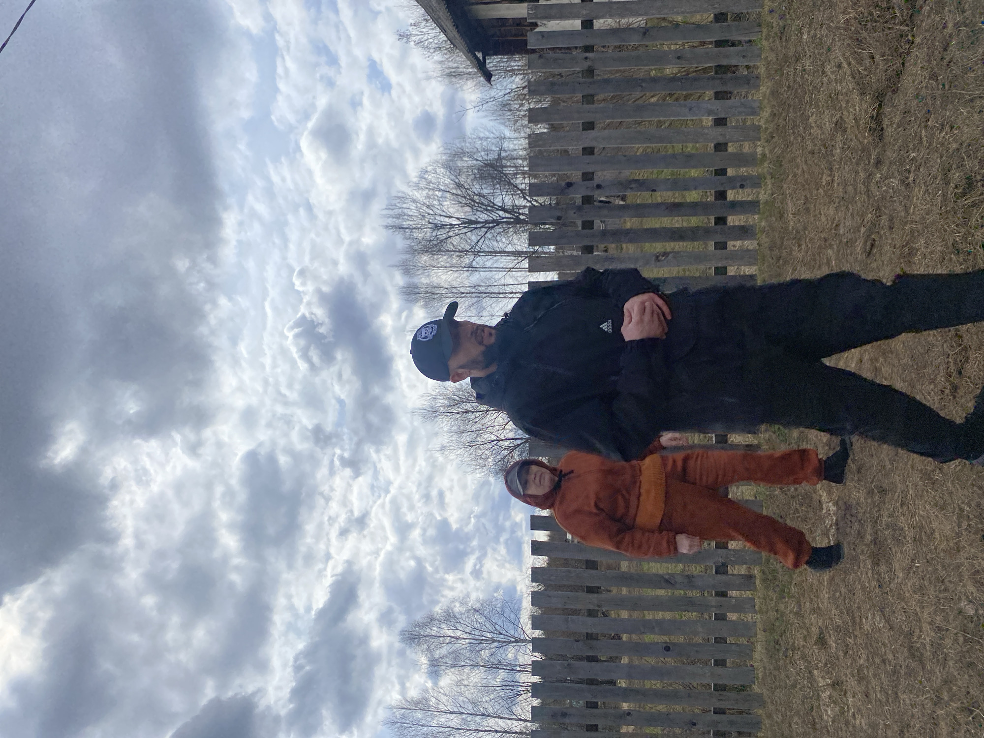 The photo appears to have been taken outdoors on a cloudy day in what seems to be a rural or semi-rural setting, possibly in Zhigalki, Russia.
A man in a black jacket, black pants, and a baseball cap stands next to a younger person wearing a rust-colored hooded jumpsuit. The backdrop includes a weathered wooden fence and dry grass. Above, the sky is filled with heavy, overcast clouds. In the distance, bare trees can be observed, suggesting it might be late fall or early spring. The quality of light and muted colors suggest a cold or overcast climate. The photo shows a casual scene, but there are no clear activities being performed other than posing for the shot.
