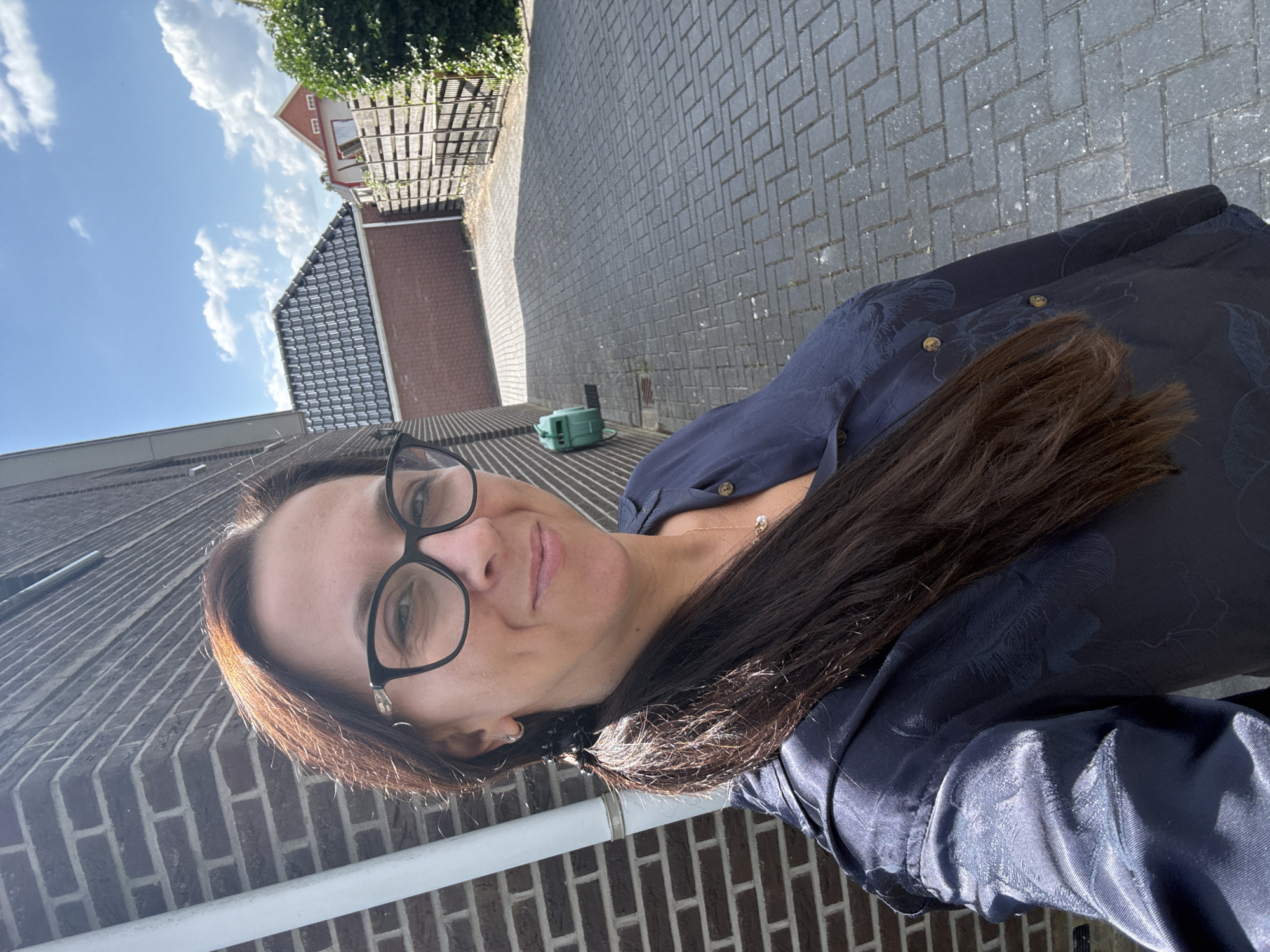 The image depicts a woman with long, dark brown hair tied in a ponytail, wearing black-rimmed glasses and a dark blue blouse with a subtle floral pattern. She is standing in an outdoor setting, likely a residential area, with a brick wall to her left and a paved walkway or driveway in front of her. The background shows houses with tiled roofs, a wooden fence, and lush green foliage. The sky is blue with scattered clouds, suggesting fair weather. The scene appears to be a selfie taken in the afternoon. The location is identified as Marum, Netherlands.