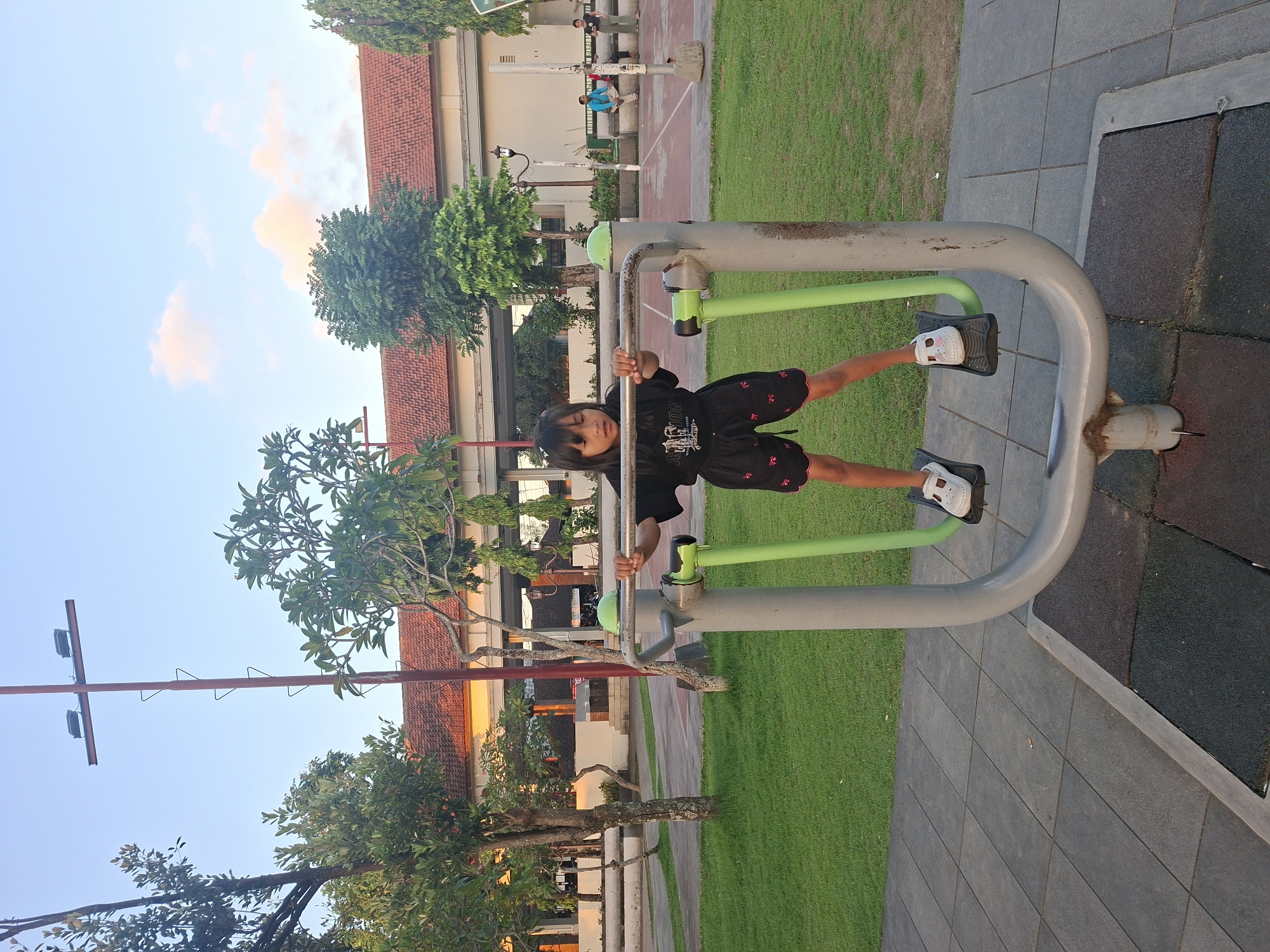 In Gianyar, Indonesia, a young girl is photographed outdoors at a public park. She stands on the footplates of a grey and green outdoor exercise machine, holding onto its handlebars. The girl wears a black t-shirt and black shorts with a red trim and embroidered pattern. She has dark hair and is wearing white sandals. The exercise equipment is situated on a paved area surrounded by a manicured lawn and trees. In the background, a building with a red-tiled roof is visible, along with a tall lamppost. The sky above is clear with a few light clouds.