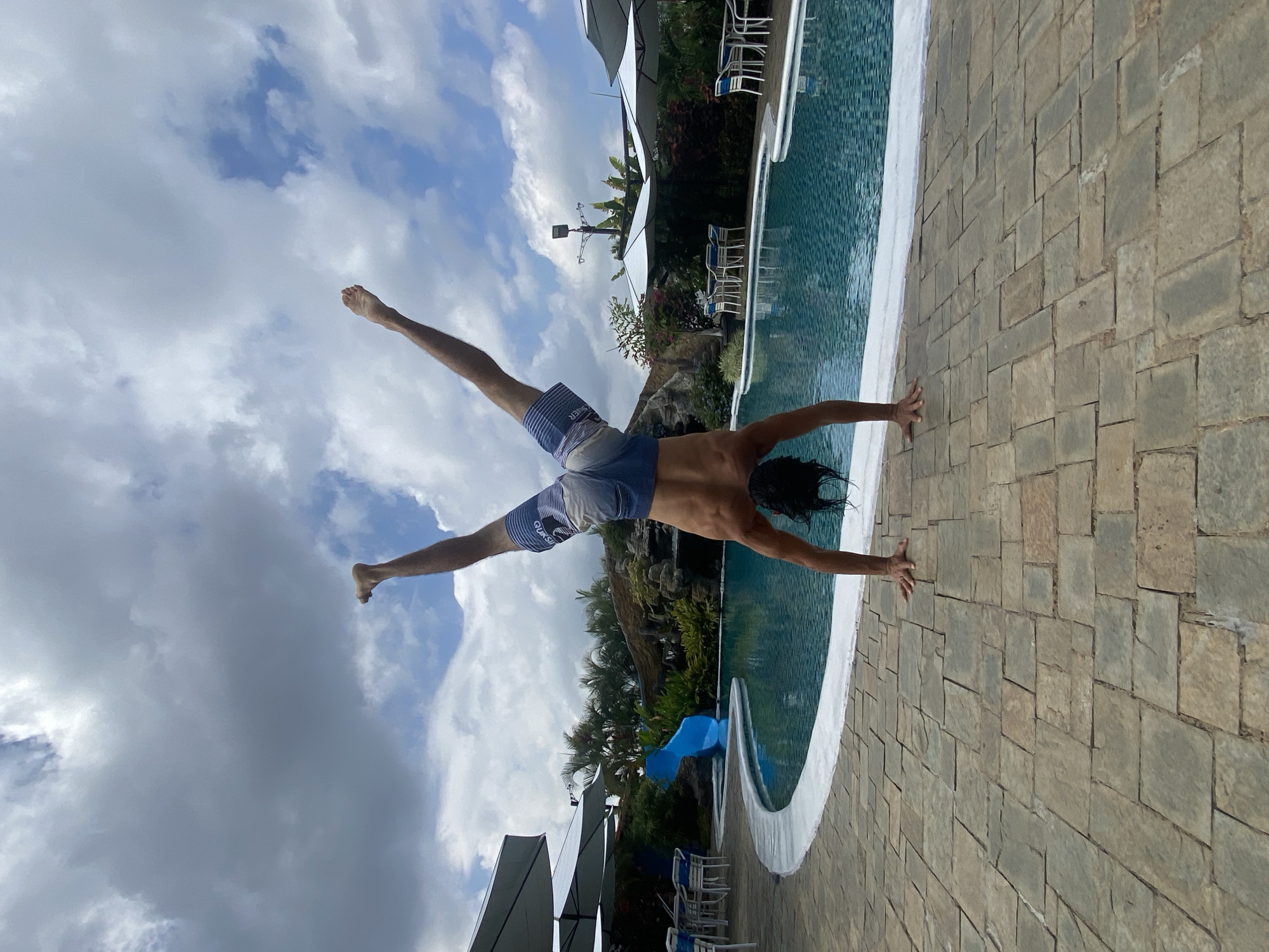 This media file depicts an active outdoor scene at a swimming pool in Vallecito, Venezuela. The central subject is a person, appearing male with dark, wet hair, captured mid-action. He is wearing blue and white patterned swim shorts, with the word "QUICKSILVER" visible on the left leg. The person is positioned upside down, with hands placed on the paved pool deck and legs splayed upwards towards the cloudy sky, suggesting either a handstand or the beginning or end of a dive into the water.
The swimming pool itself features clear, blue water and a clean white coping along its edge. The pool deck is constructed from light brown and grey rectangular pavers. In the background, a lush tropical environment is visible, with abundant green foliage, including palm trees and other dense vegetation. Several large white patio umbrellas and white patio chairs with blue accents are also part of the scene, indicating a resort or recreational area.
The time of day appears to be bright daytime, likely late morning or early afternoon, under a partly cloudy sky with visible patches of blue. The image conveys a sense of athleticism and leisure in a vibrant, tropical setting.
