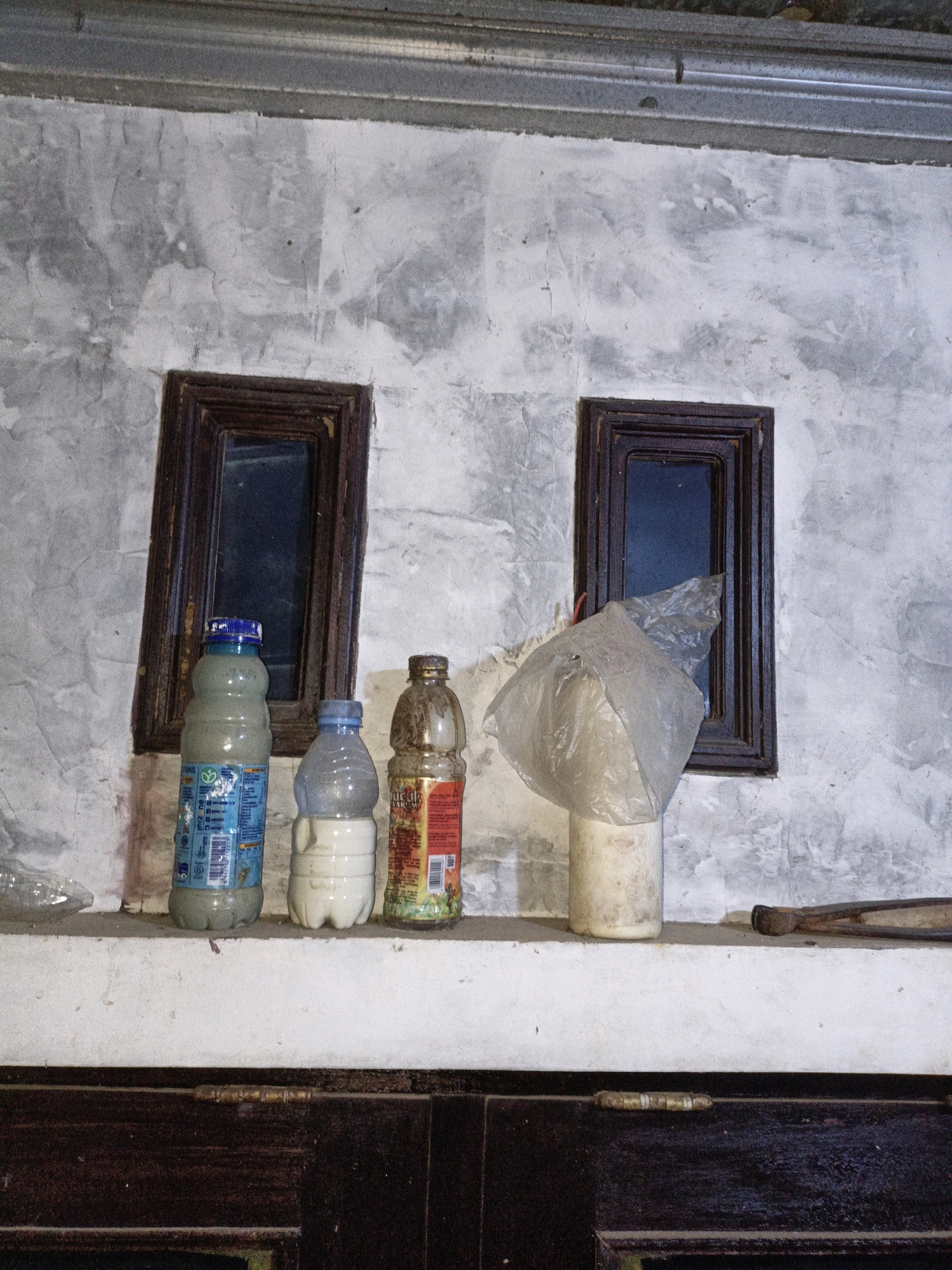 In Cikal, Indonesia, on a white shelf in front of a textured grey wall with two small, dark-framed windows, are several objects. To the left, a clear plastic bottle lies on its side. Next to it, standing upright, is a blue and white bottle containing a grey liquid. To its right, a white bottle holds a lighter colored liquid. Further to the right, a brown bottle with a red and yellow label contains a dark liquid. To the right of the brown bottle, a white cylindrical object is partially covered by a crumpled clear plastic bag. A dark-colored tool with metal ends rests on the shelf to the far right. Below the shelf, a dark wooden surface with visible hinges is present. The lighting suggests an indoor environment, likely a storage area.