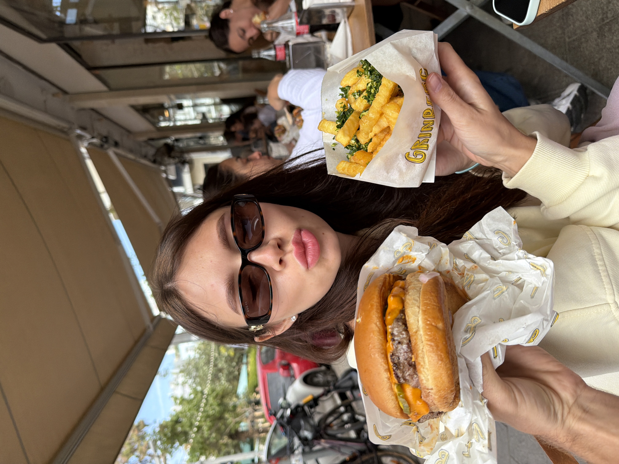 The scene captures a young woman dining outdoors in Tel-Aviv, Israel. She holds a burger in her right hand and fries in her left. Other patrons are seated at nearby tables. The backdrop includes buildings, trees, cars, and bicycles.