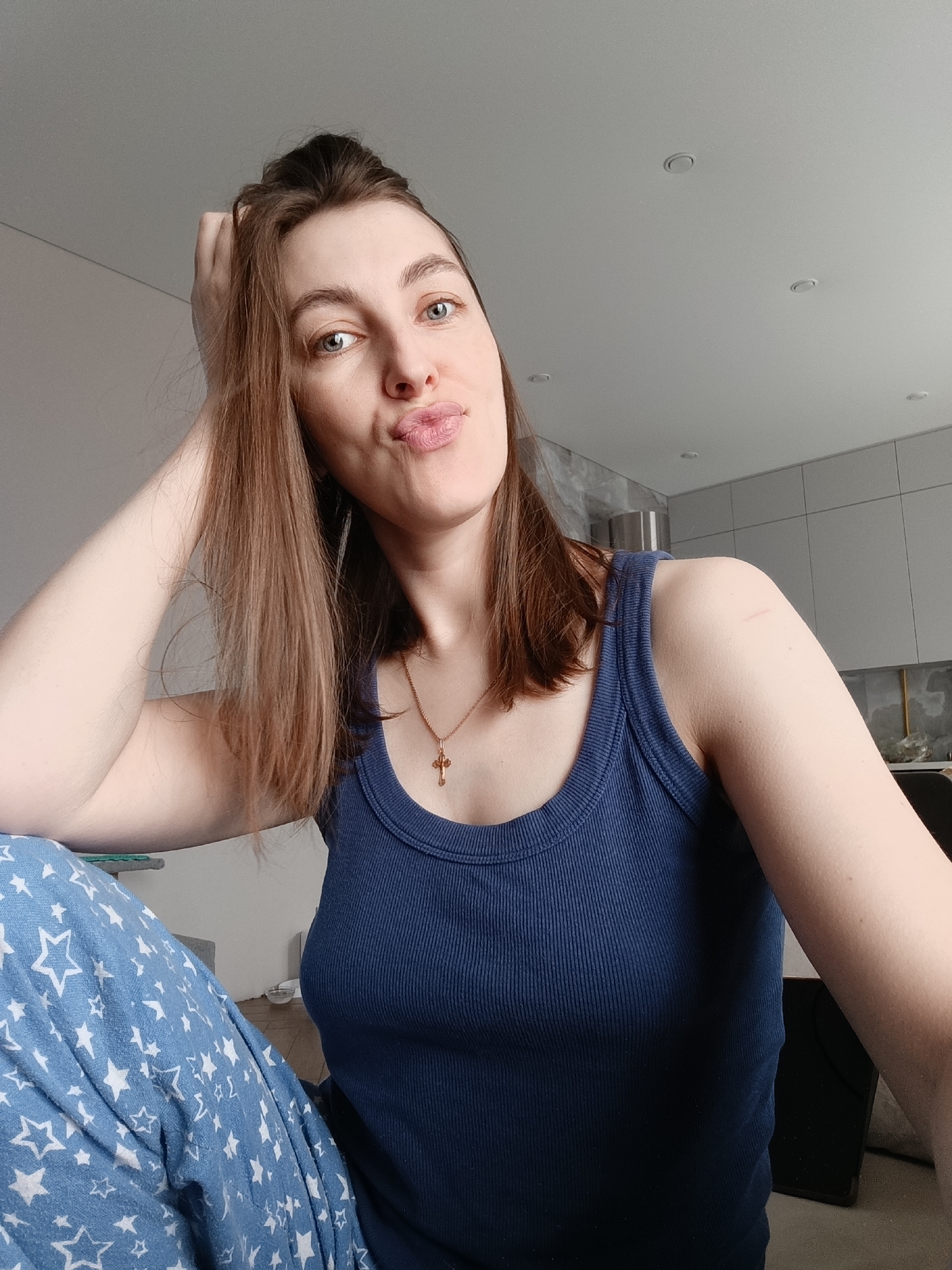 A woman is captured in a close-up selfie, looking directly at the camera with a playful, puckered-lip expression. Her head is tilted slightly, and her long, dark brown hair falls over her shoulders. Her right hand rests casually on the back of her head, with fingers visible in her hair. She has light blue or grey eyes.
She is dressed in a navy blue, ribbed tank top, and blue pajama bottoms with a white star pattern are visible on the lower left. A delicate gold chain with a small gold crucifix pendant hangs around her neck. A small, light-colored scar is noticeable on her left shoulder.
The background reveals a modern indoor setting. A light grey ceiling with multiple recessed circular lights is visible in the upper portion of the frame. To her right and behind her, sleek, light grey, flat-panel kitchen cabinets are present, suggesting a contemporary kitchen or living area. A shiny object, possibly a glass vase, rests on a counter, and a dark rectangular object (perhaps a tablet or screen) is partially visible on the far right. The ambient lighting suggests it is daytime. The woman's relaxed posture and playful expression convey a casual mood. No text is visible in the image.