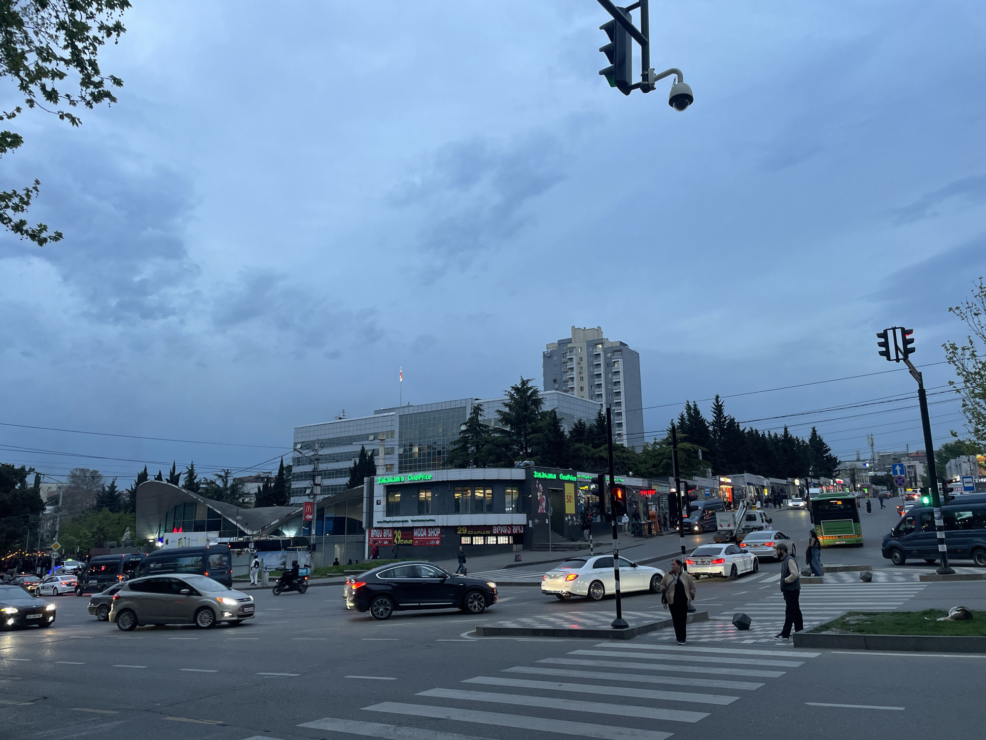 This image depicts a bustling city intersection in Tbilisi, Georgia, captured during dusk or early evening under an overcast sky. The scene is active with both vehicular and pedestrian traffic.
In the foreground, a prominent crosswalk spans the street, where several pedestrians are either walking or waiting. Various cars are on the road, including a silver Ford Focus, a black BMW X4, a white Mercedes-Benz E-Class, and other vehicles, some with their headlights on. Traffic lights are visible, with one showing a green signal to the right and another displaying red to the far right. A security camera is mounted on a pole above the traffic signals.
The background features commercial buildings and residential structures. A distinctive building with a curved, possibly metal, roof is visible on the left, while a taller, more generic apartment building rises in the distance on the right. Shopfronts along the street are illuminated, showcasing various businesses. Trees and some sparse greenery are present throughout the urban setting. A green municipal bus and a dark blue van with "POLICE" written on it are also part of the traffic.
Visible text includes "33636300 OnePrice" on a larger building and "OnePrice" again on a smaller shop below it. A sign also reads "ames ama 20 MODA SHOP" and "29 ლარად მოდა შოპ" (which translates to "for 29 Lari Moda Shop," Lari being the Georgian currency), indicating a clothing store. A "50%" discount sign is also noticeable.