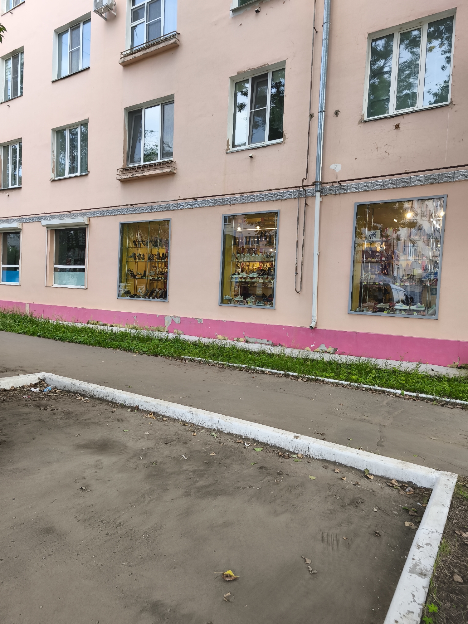 The image shows the exterior of a pink multi-story building with several windows. On the ground floor, there are three large display windows showcasing shoes. The windows are brightly lit from within, and the shoes are displayed on shelves. The building is located in Komsomolsk-on-Amur, Russia. In front of the building, there is a sidewalk and a patch of grass. The ground in the foreground is a dirt or asphalt surface with some scattered leaves and debris.