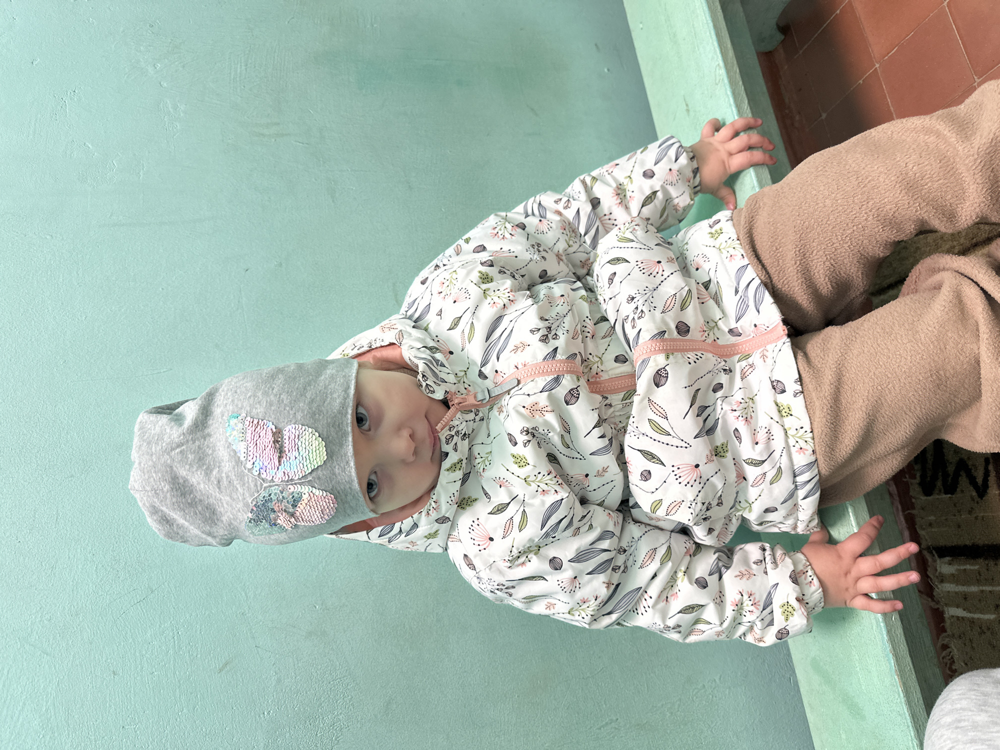 A baby wearing a gray hat with a sequined butterfly appliqué, a white floral patterned jacket with a light pink zipper and tan sweatpants is sitting by a pale green wall, possibly in Прикамский, Russia. One hand is holding onto the top of the wall and one hand is holding onto the side. The floor is a mottled brown.