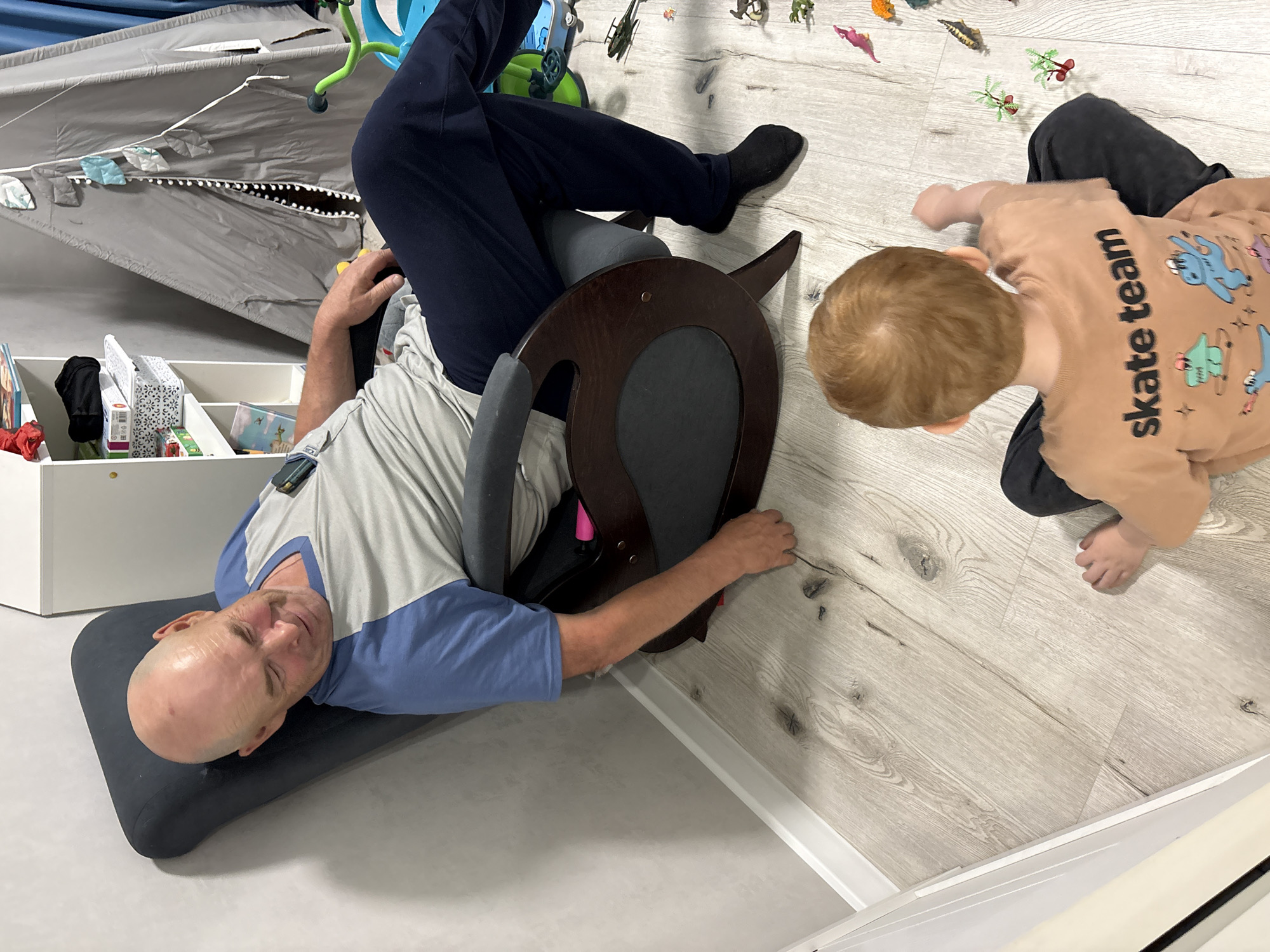 A man is reclined backward on a dark grey support, with his legs elevated up the back of a dark wood kneeling chair. The man is wearing dark blue trousers, a grey and blue shirt, and black socks. His head rests on a dark grey pillow. Next to him, a child in a light brown sweater with "skate team" printed on it is playing with a collection of toy dinosaurs. The floor is made of light wood panels. A white shelf with various objects is visible in the background. In the top-left corner of the image, part of a grey play yard can be seen. The location is likely indoors. This image could be from Izhevsk, Russia, as requested.