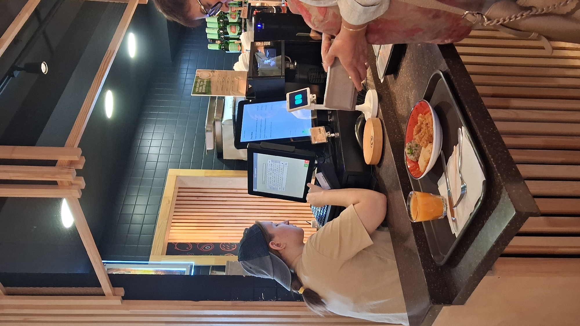 A young woman wearing a tan t-shirt and a blue baseball cap is operating a cash register, facing two computer monitors. One monitor displays a keyboard layout, and the other shows a menu with Russian text. She is seated behind a dark countertop. On the countertop, a tray holds a glass of orange juice, a small bowl with food, and cutlery. To the right, another person, partially visible from the shoulders up, is wearing glasses and holding a gray wallet, possibly interacting with a payment terminal. Behind them, shelves are stocked with bottles. The background features wooden vertical slats and black tiled walls, illuminated by overhead lights. The scene appears to be a food service counter in a cafe or restaurant.