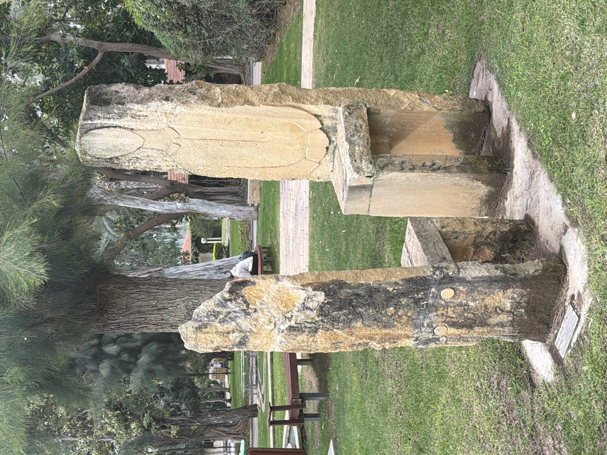 This image depicts a stone monument in what appears to be a park-like setting in Herzliya, Israel. The monument consists of two large stone pieces, seemingly carved with symbolic designs. The upper stone features a carved, oval shape and intricate detailing. The lower stone, darker in tone, has a rougher texture. The monument is set on a stone base surrounded by green grass. Trees are visible in the background, along with benches and a path. The overall environment suggests a peaceful public space within the city of Herzliya.