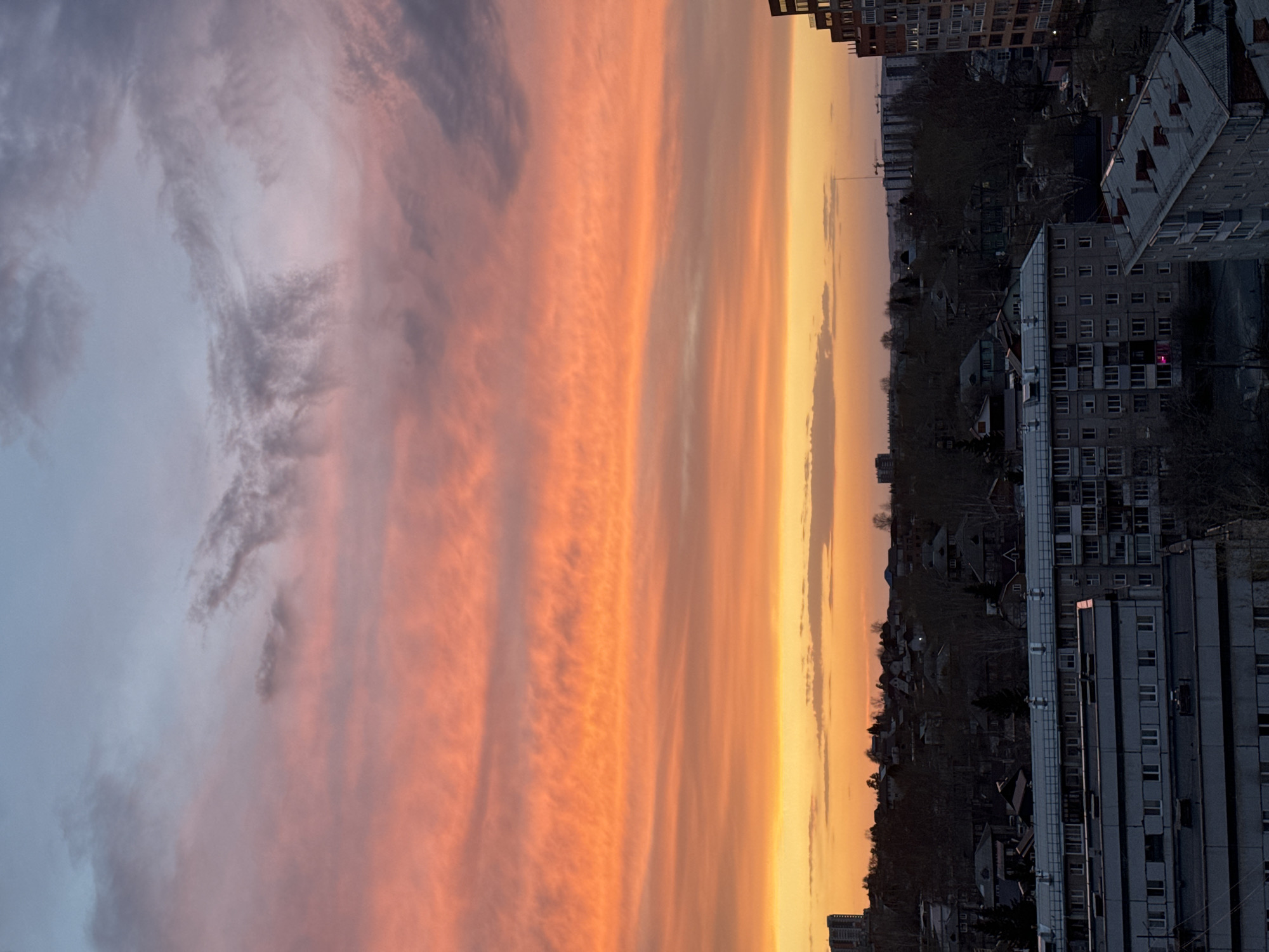 The image shows a sunset over Krasnoyarsk, Russia, with buildings and trees silhouetted against the sky. The sky is predominantly orange and yellow near the horizon, transitioning to shades of pink and grey higher up. There are several high-rise apartment buildings and other structures visible. The sky has layers of clouds.
