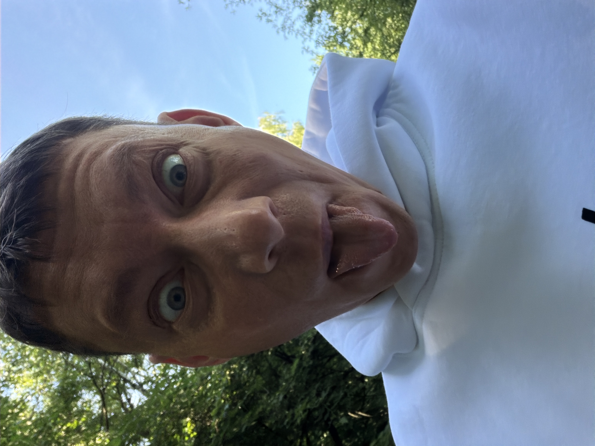 A close-up, selfie-style photograph captures a Caucasian male with short brown hair, protruding his tongue with wide, surprised-looking eyes. He is wearing a white hooded sweatshirt. The background consists of lush green trees and a bright blue sky, suggesting an outdoor setting, possibly a park or wooded area. There is no visible indication of the location being in Moscow, Russia.