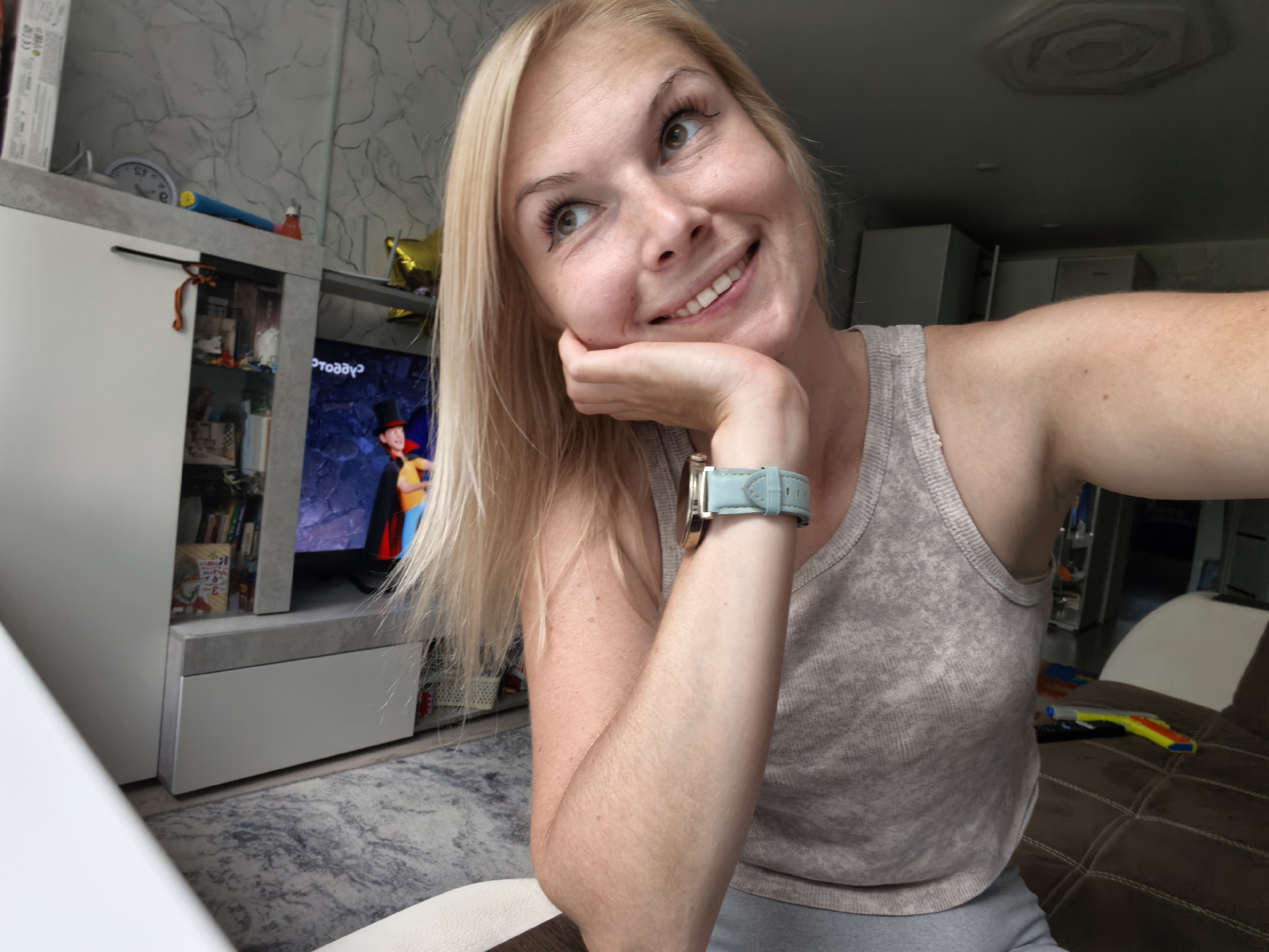 The image depicts a blonde woman with a light complexion, freckles, and green eyes, wearing a grey tank top and a light blue watch. She is indoors, resting her chin on her hand and looking upwards and to the left, with a wide smile. A television screen displaying a cartoon character in a top hat is visible in the background, along with a grey and white entertainment center filled with books and other items. The room appears to be a living area, with a patterned rug on the floor and a dark brown sofa in the foreground. The lighting suggests it is daytime. The provided context mentions Sterlitamak, Russia, but the image itself does not contain any overt visual cues to confirm or deny this specific location.