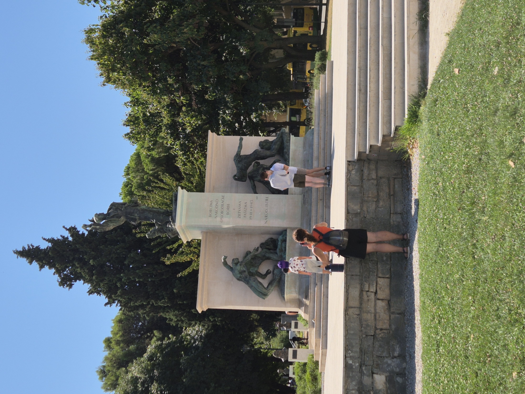 In the city of Grad Pula, Croatia, a monument stands prominently in a public space on a sunny day. The monument features bronze sculptures depicting struggle and sacrifice, set against a stone backdrop with inscribed text. At the base of the monument, a woman with a backpack is taking a photo of the scene with her phone. Nearby, a child in a purple hat observes the monument. Two men stand together, embracing, to the right of the monument. In the background, a row of trees forms a dense green canopy, and yellow buses are visible on a street beyond the monument. The ground in front of the monument is covered in green grass and stone pathways.