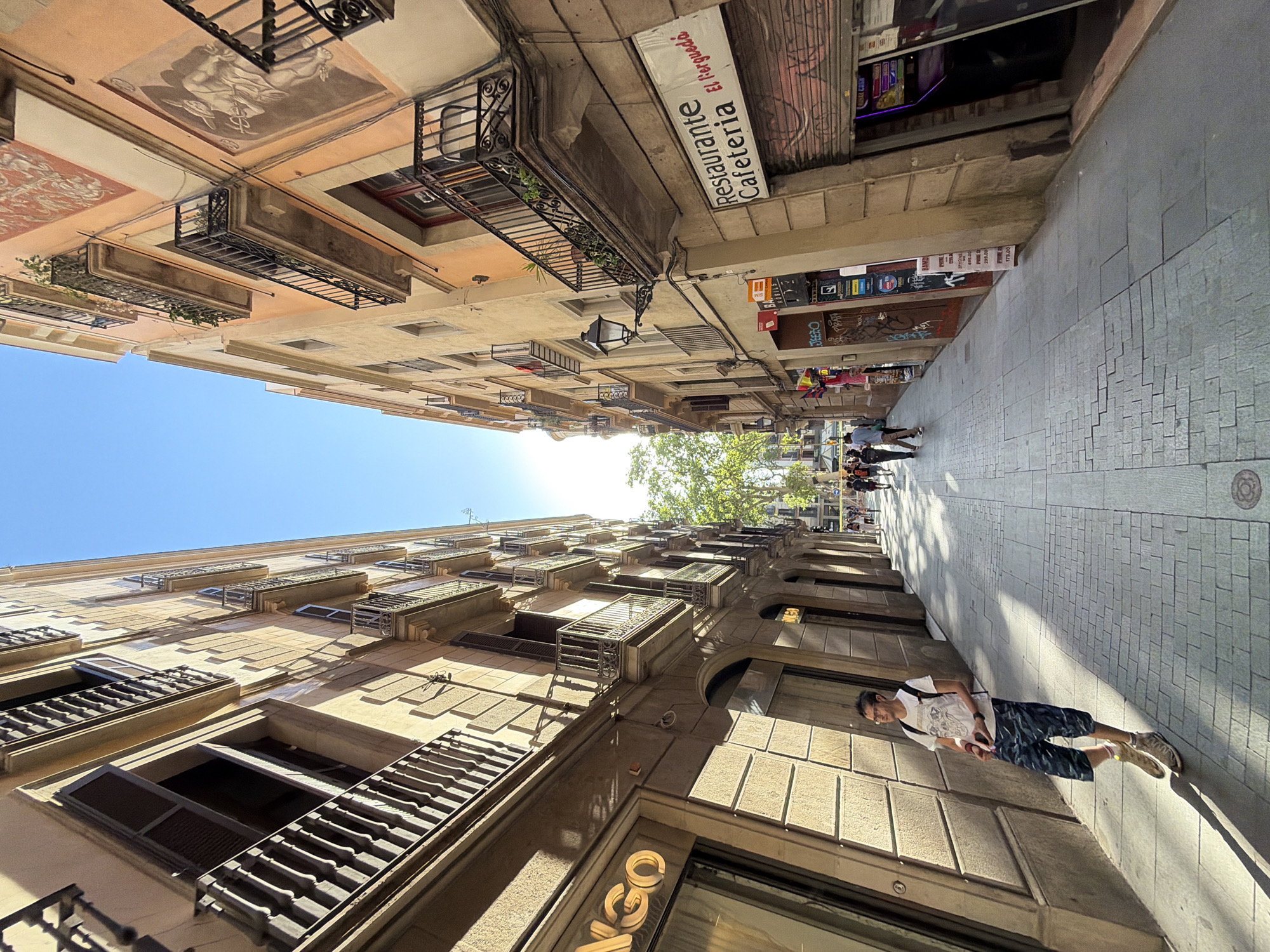 The image depicts a street scene in Barcelona, Spain. Two tall buildings with ornate balconies and shuttered windows face each other, creating a narrow, shaded alleyway. The buildings are constructed of light-colored stone, with some displaying faded murals. The sky is a clear blue, indicating fair weather. The time of day appears to be midday, given the strong shadows cast by the buildings and the bright sunlight.
In the foreground, a young man wearing a white t-shirt and patterned shorts is leaning against the building on the right. He is looking towards the camera and appears relaxed. Further down the street, a few other people can be seen walking, suggesting a moderate level of pedestrian traffic.
Signs are visible on the buildings. One sign on the left building reads "Restaurante Cafeteria El Vergudo." Another sign on the right building, though partially obscured, appears to be a price list for what looks like wine. The architecture, cobblestone pavement, and the presence of a "Restaurante" sign all contribute to the Spanish setting. The overall mood of the image is one of urban exploration and everyday life.