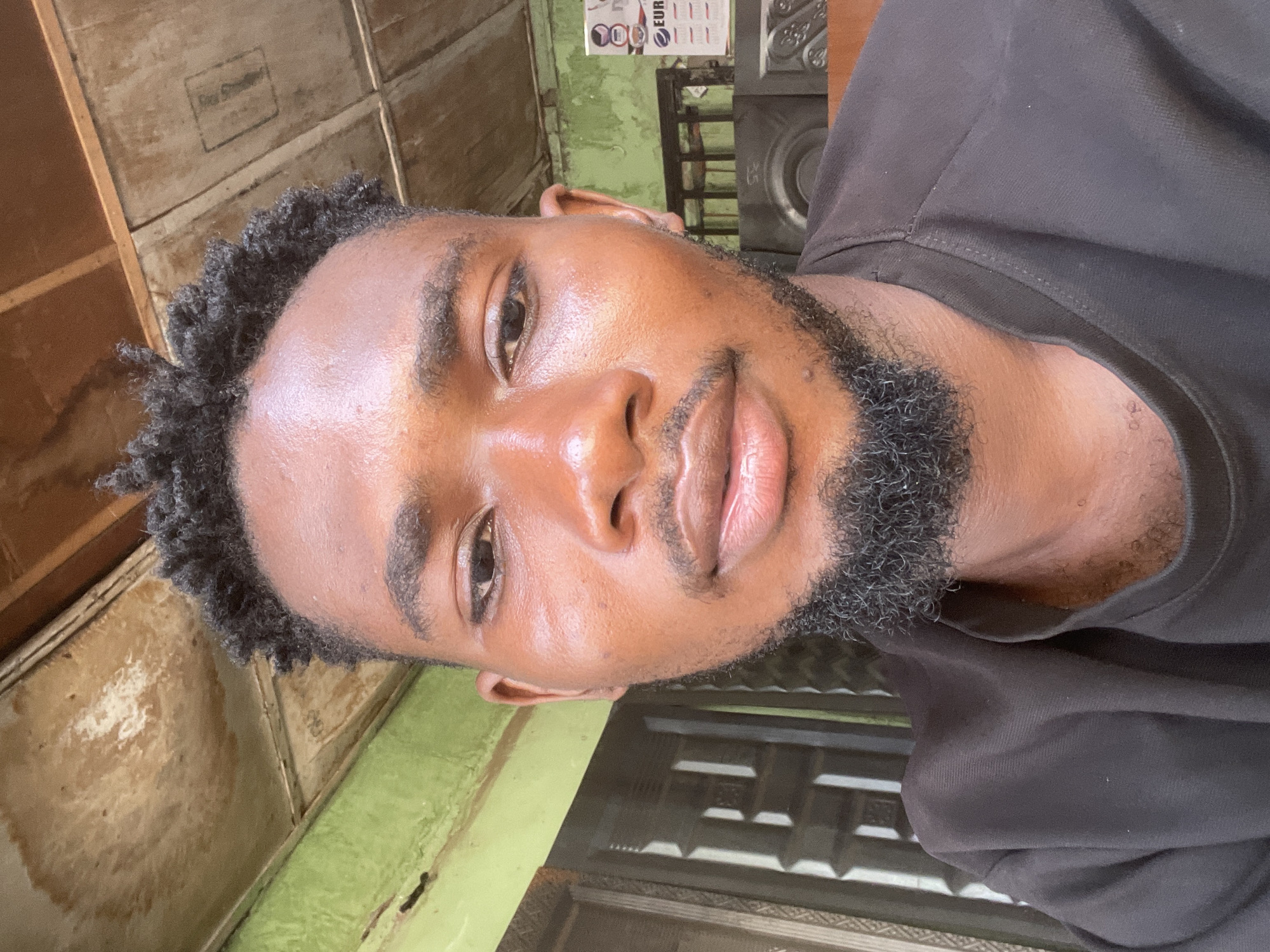 A close-up headshot shows a Black man with short, dark, curly hair and a dark beard. He is wearing a black shirt. The background consists of a wooden wall painted in a faded beige and green, with a window frame on the right and a calendar hanging on the wall. The location is likely Chikaji, Nigeria.