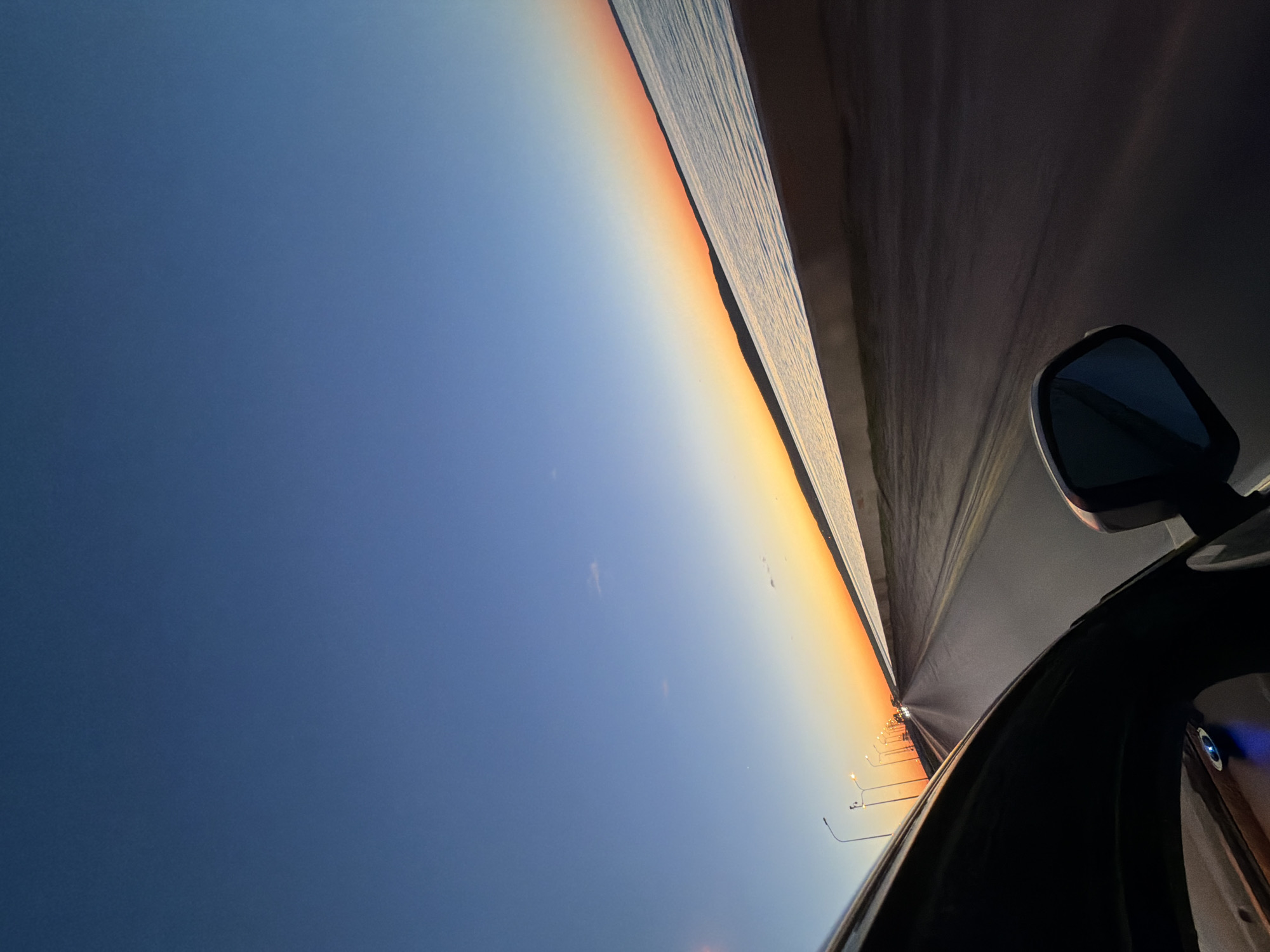 The image shows a view from inside a vehicle moving along a road, with the road and sky visible. The right side of the image features part of the vehicle interior, including the side mirror. The road ahead appears to be lined with street lights. The sky transitions from a dark blue at the top to orange near the horizon, suggesting it could be sunset or sunrise. While the exact location within Chaikovsky, Russia, cannot be determined from the image, the presence of a road and street lights indicates an urban or suburban environment. There are no people visible in the image.