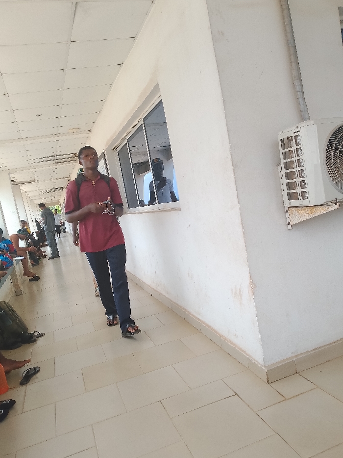 The image shows a hallway in what appears to be a building in Umueshi, Nigeria. The hallway has a tiled floor and a white wall with a window and an air conditioning unit. People are sitting and standing in the hallway, with one man walking toward the camera. He is wearing a red shirt, dark pants, and sandals, and has a backpack on. A cross is visible on his chest.