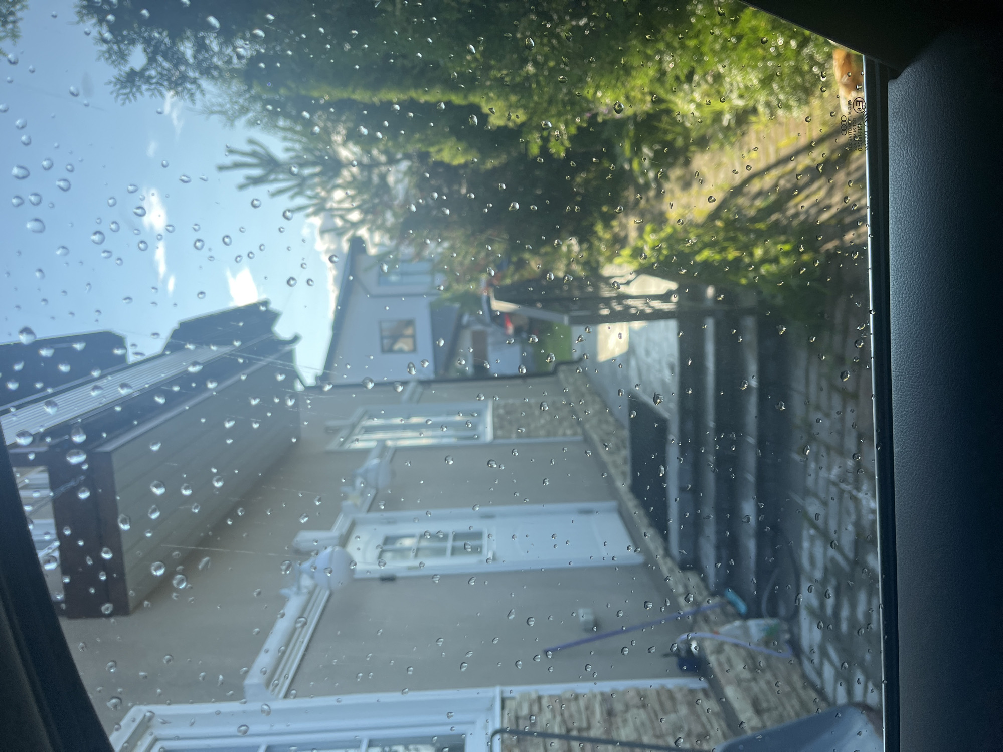 The image, taken from inside a vehicle looking out through a rain-streaked window, depicts a residential area. In the foreground, the lower part of the car's interior is visible. The view through the window shows the exterior of a house with a beige facade and a white door. Steps lead up to the entrance. To the right of the house, there is a path bordered by lush green foliage and trees. The sky above is blue with scattered clouds, and sunlight filters through, casting shadows. The presence of water droplets on the window suggests recent or ongoing rainfall. The location is specified as Vladimirovka, Russia.