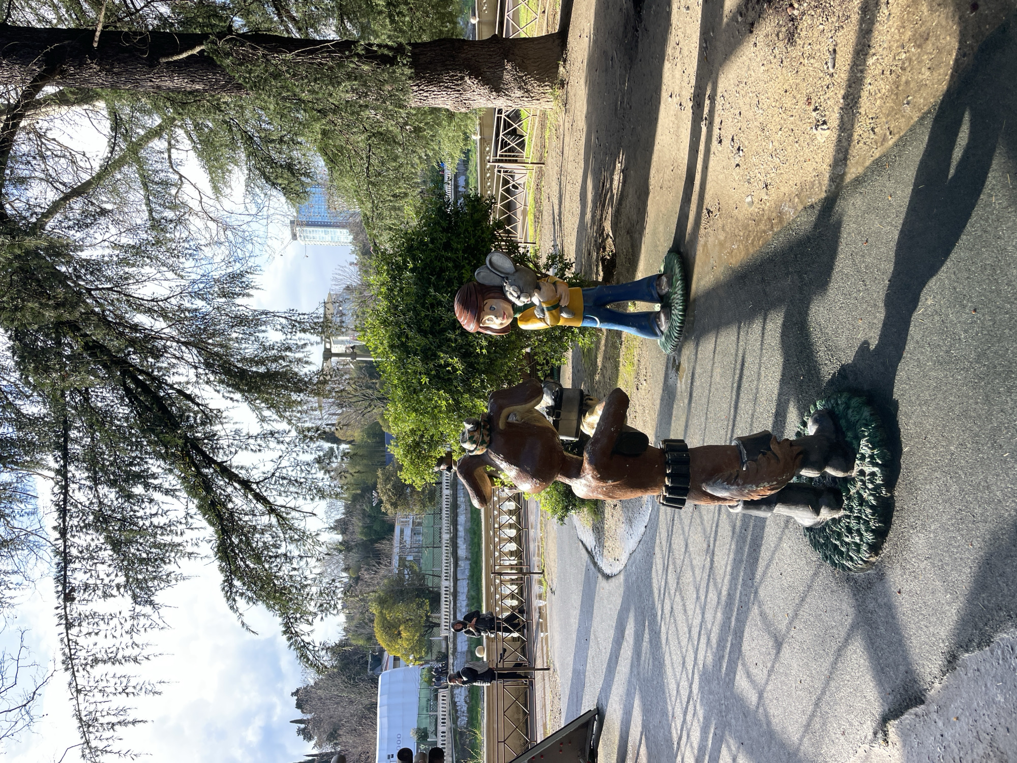 The image shows a sunny day in Sochi, Russia, likely in a park or recreational area. The scene features two painted statues: one of a child holding a toy rabbit and the other of a cartoon dog. The sculptures are positioned on a paved surface near a tree and a metal railing, with greenery in the background. In the distance, buildings are visible, including a high-rise structure. There are shadows cast by the trees and railing on the ground.