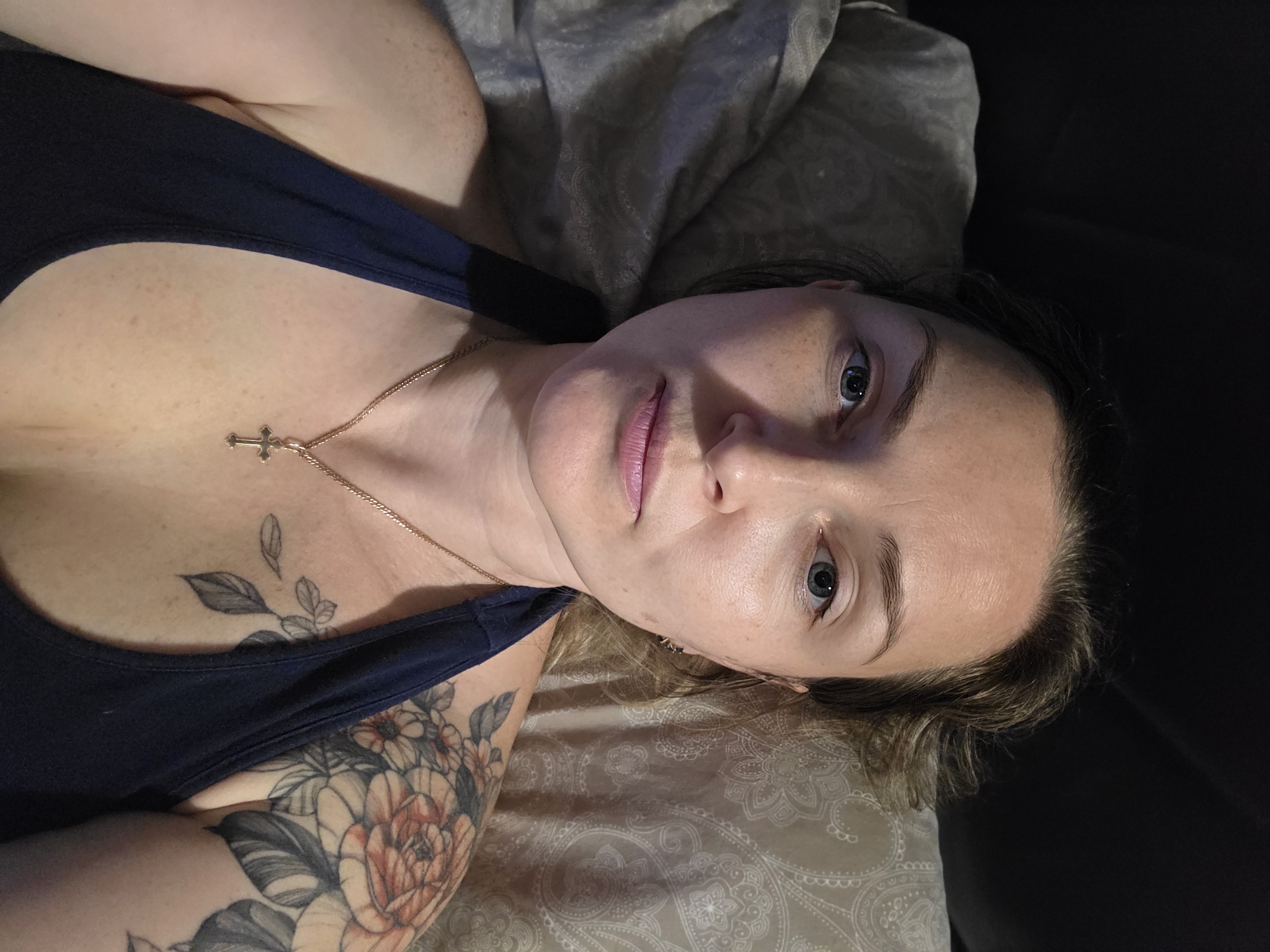 An adult woman is depicted lying down indoors, looking directly at the camera, in what appears to be an evening or nighttime setting in Chaikovsky, Russia. She has light-colored eyes and fair skin, with visible freckles across her chest and face. Her lips are a soft pink. She wears a dark blue tank top or camisole, revealing a gold chain necklace with a small gold cross pendant resting on her chest.
Notable tattoos adorn her body: a large, vibrant floral design with peach-toned flowers and dark leaves covers her left upper arm and shoulder, while smaller, monochromatic leaf or vine tattoos are visible on her upper chest near her collarbone. Her hair, light brown or blonde, is casually styled around her head.
The woman is resting on a pillow with a subtle, light-colored patterned fabric, possibly a paisley design, suggesting she is on a bed or couch. The right side of the image (to her left) is dark, indicating a shadowed area or part of the furniture. The lighting is soft and somewhat dim, illuminating her face and upper body from an unseen source, creating a calm and intimate atmosphere. Her expression is neutral and direct. The photograph is composed from a slightly elevated and tilted angle, giving a diagonal orientation to the subject. No text is visible in the image.