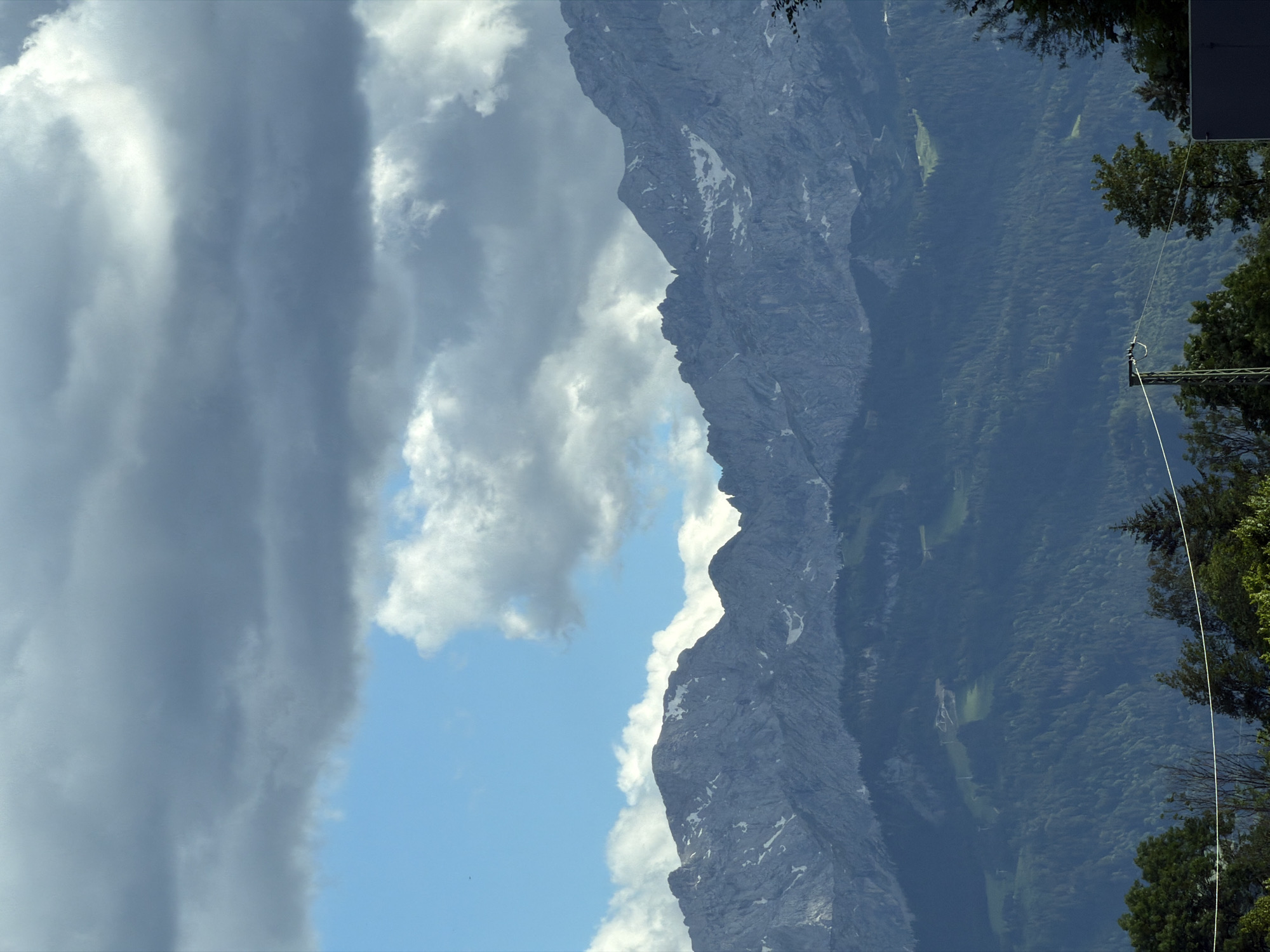 The image depicts a panoramic view of a mountainous landscape, likely in the Bavarian Alps region near Oberau, Germany, given the terrain and architecture. In the foreground, lush green trees and foliage line the bottom of the frame, with a stark white cable running horizontally across the middle distance. Beyond the trees, a series of imposing mountain ranges dominate the scene, their rugged slopes dotted with patches of snow and scree. The mountains are covered in a dense forest of dark green conifers in the lower and mid-sections, transitioning to rocky, bare peaks at higher elevations. The sky above is a dramatic mix of deep gray, heavy clouds and patches of bright blue sky, with scattered white clouds adding texture and depth. The lighting suggests a partly cloudy day, with the sun intermittently breaking through. In the far distance, nestled within the mountains, hints of human activity are visible, possibly including ski lifts or mountain trails, though they are not distinctly clear. The overall impression is one of natural grandeur and a vast, untamed wilderness.