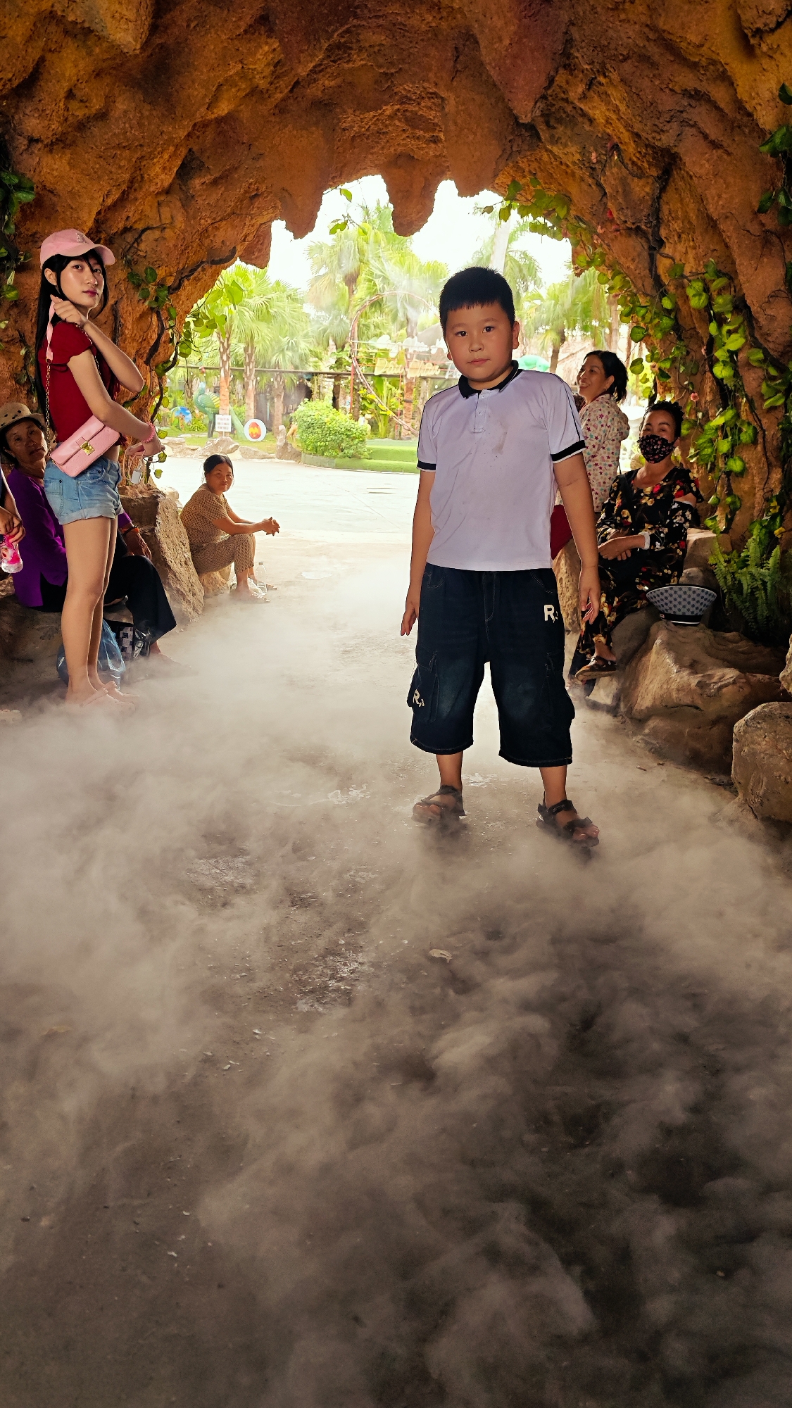 A young boy in a white polo shirt and dark shorts stands in the foreground of a misty, cave-like setting. He is looking directly at the camera, with his hands by his sides. To the left, a young woman in a pink cap and red top, wearing denim shorts, is looking away from the camera and posing. Behind her, several people are seated on rocks, some of whom are obscured by mist. The environment appears to be an outdoor attraction, possibly a theme park or garden, with lush greenery and palm trees visible in the background. The ground is covered in a thick, white mist, creating a hazy atmosphere. The overall scene suggests a playful or recreational setting.