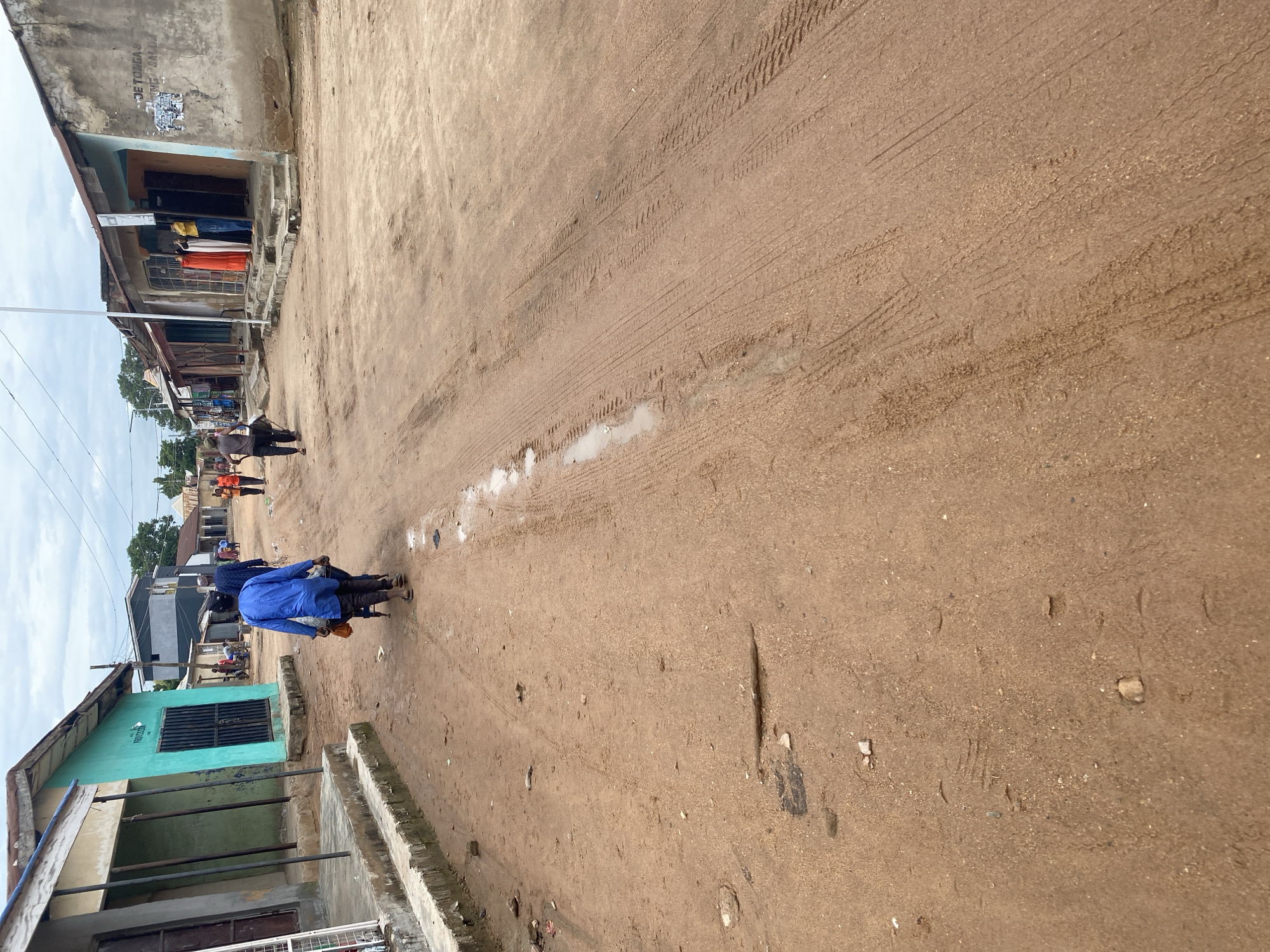 The image depicts a street scene in Jalingo, Nigeria, likely during the daytime given the bright sky and general illumination. The street is unpaved, composed of dirt and sand, with visible tire tracks and some small puddles, suggesting recent rain or a naturally damp area.
In the foreground, a person wearing a bright blue shirt is riding a motorcycle, facing away from the camera. Behind them, several other individuals are visible on foot and possibly also on motorcycles, further down the street. On the left side of the street, there are buildings that appear to be shops or houses. One building in the immediate left foreground is painted turquoise and has a barred window. Further down, another building has an open doorway where several people are standing, with colorful garments, possibly clothing for sale, displayed.
The architecture is simple, reflecting a local style. The overall atmosphere appears to be that of a typical, active day in a Nigerian town. There are no explicit indications of a specific event, but rather a snapshot of daily life and movement. The weather seems overcast, contributing to the soft lighting of the scene. The ground is uneven and shows signs of wear and tear, typical of an unpaved road in a populated area. There are some small pieces of debris scattered on the ground. No discernible text is visible in the image.