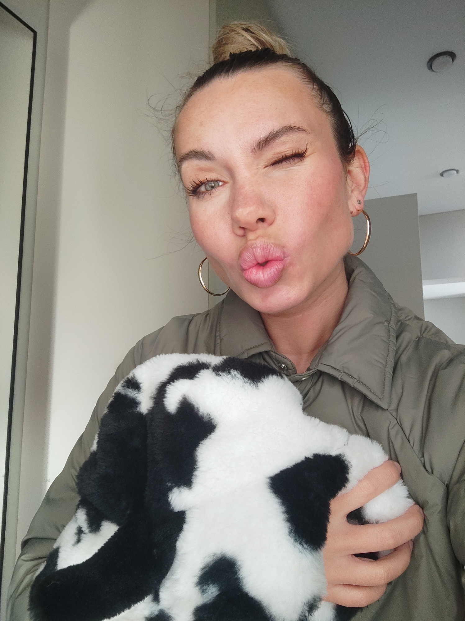 A woman is taking a selfie indoors, holding a black and white cow-patterned, fluffy object. She has her hair in a high bun, is wearing gold hoop earrings, and a green jacket. She is winking and pursing her lips. The background includes a white wall and a door. There is no evidence of Votkinsk, Russia.