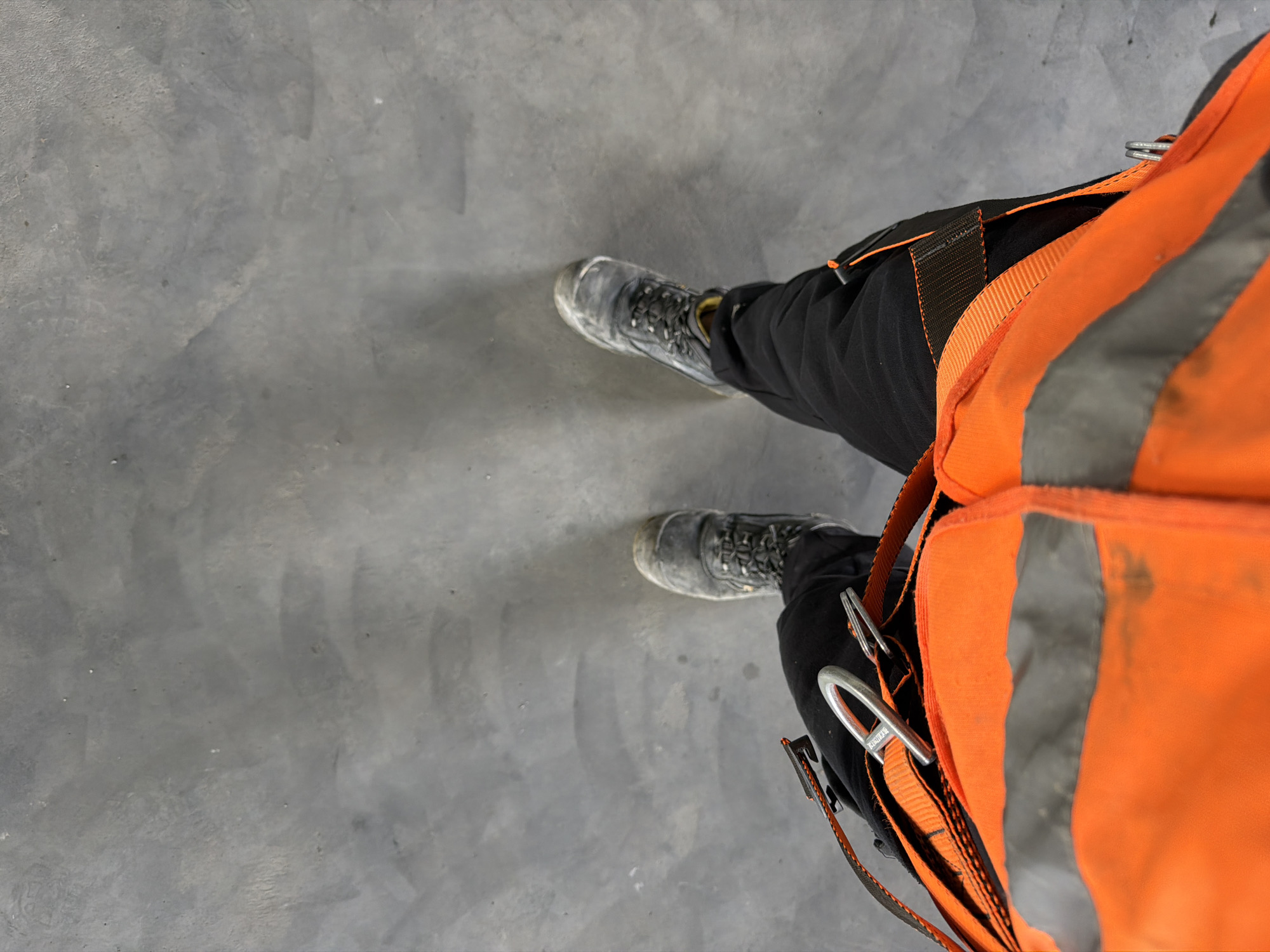 The image is a first-person perspective shot of a person standing on a grey concrete surface. The person is wearing black work pants and a high-visibility orange vest with reflective gray stripes. They are also wearing dark work boots. There is a safety harness with metal clips visible on the vest and pants. The concrete surface appears to be a floor or ground within an urban or industrial setting. The location is identified as Stróża, Poland.